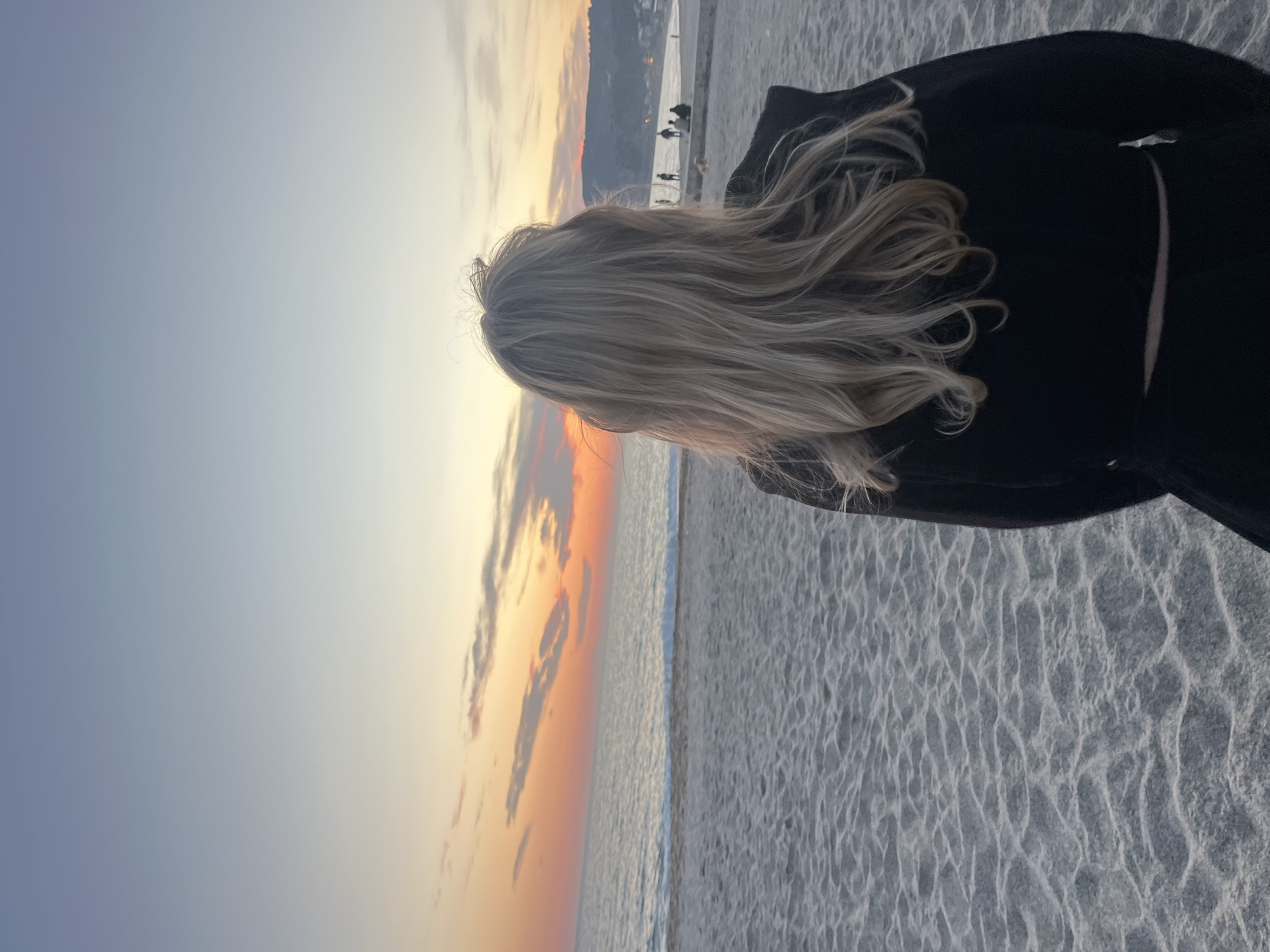 The image depicts a serene sunset scene on a beach, viewed from a slightly elevated perspective behind a person. The photograph is oriented vertically (portrait mode).
In the foreground, the back of a person with long, wavy blonde hair is visible, turned towards the ocean. They are wearing a dark, possibly black, coat or sweater, the fabric of which is visible over their right shoulder and back. The person stands on a sandy beach, characterized by subtle ripples and textures.
The mid-ground features the calm waters of the sea, meeting the shore with a gentle line. The horizon is prominent, dividing the sea from the sky.
The sky dominates the upper and left portions of the image (when viewed upright), displaying a beautiful gradient of colors characteristic of sunset. The sky transitions from a soft light blue at the top, through warmer hues of orange and yellow near the horizon, with scattered clouds reflecting these warm tones.
In the distant background, along the coastline, there are faint outlines of mountains or hills. Further to the right, a structure resembling a pier or boardwalk extends into the water, with several small, silhouetted figures of people visible on it, enjoying the evening. This suggests a populated coastal area, consistent with Alanya, Turkey.
The overall activity is observation; the person is looking out at the sunset over the sea. The atmosphere is peaceful and contemplative. The time of day is clearly sunset, as indicated by the vibrant sky colors. There is no visible text in the image.