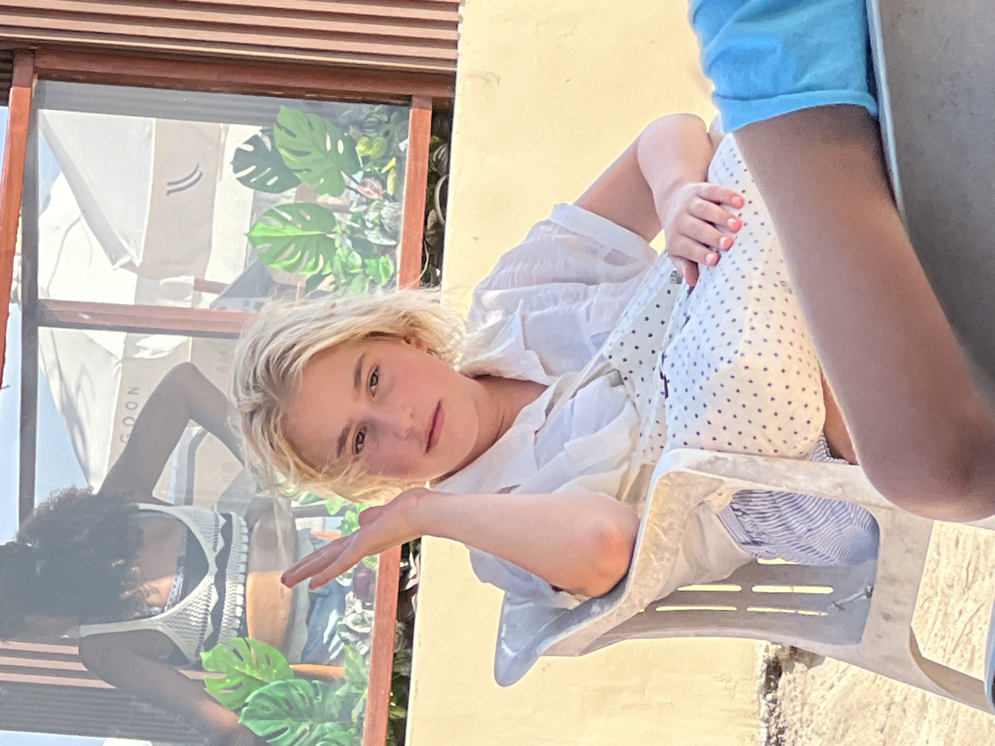 A blonde-haired girl sits in a plastic chair on a sandy beach, her left arm raised and hand in the air, looking towards the camera. She wears a white collared shirt and striped shorts. A person in a blue shirt is visible in the foreground. Behind the girl is a window with a view of umbrellas and tropical plants, suggesting an outdoor seating area at a beachside establishment in Kuta, Indonesia.