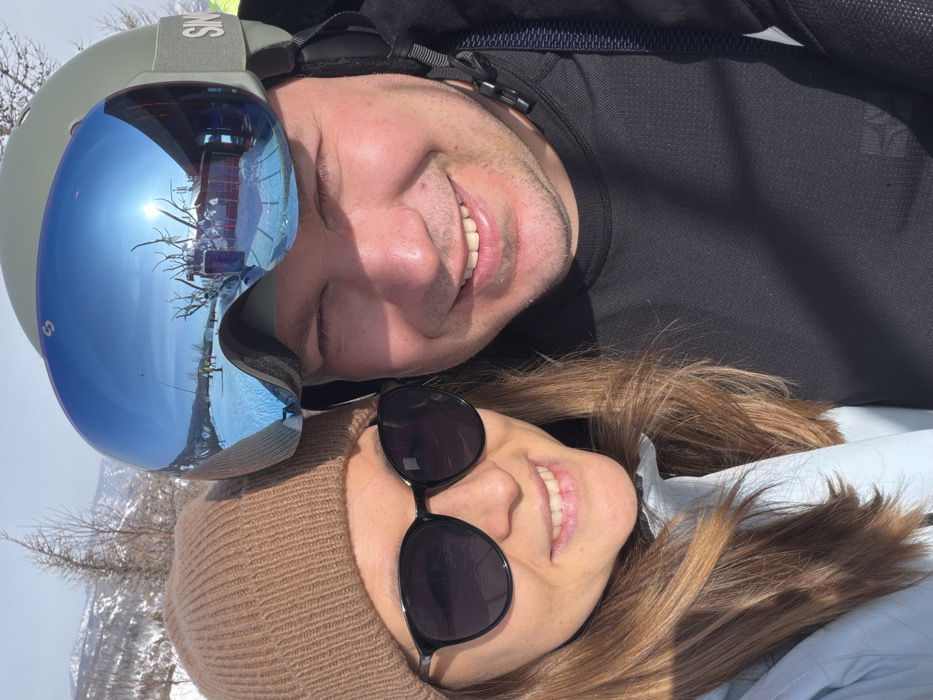 The image presents two smiling individuals, a female and a male, captured in a close-up portrait within a sunny, snow-covered mountain environment identified as Zelenaya Polyana, Russia.
The female, positioned on the left, wears a light brown knitted beanie, dark-tinted, black-framed sunglasses, and a light blue jacket or outer layer. Her light brown hair is visible beneath her beanie and around her shoulders. She is smiling broadly.
The male, positioned on the right, wears an olive green ski helmet with a visible "SIN" logo on its strap, and large blue mirrored ski goggles. The goggles reflect a detailed panoramic view of a ski resort under bright sunlight, including a snow-covered slope with discernible ski tracks, several distant figures, ski lift infrastructure, a building featuring dark windows and decorative elements resembling antlers, and bare trees against snow-capped mountains. He is wearing a black long-sleeved athletic top and is also smiling broadly. Both individuals are situated in close proximity, suggesting a shared activity or relationship in the snowy setting.
