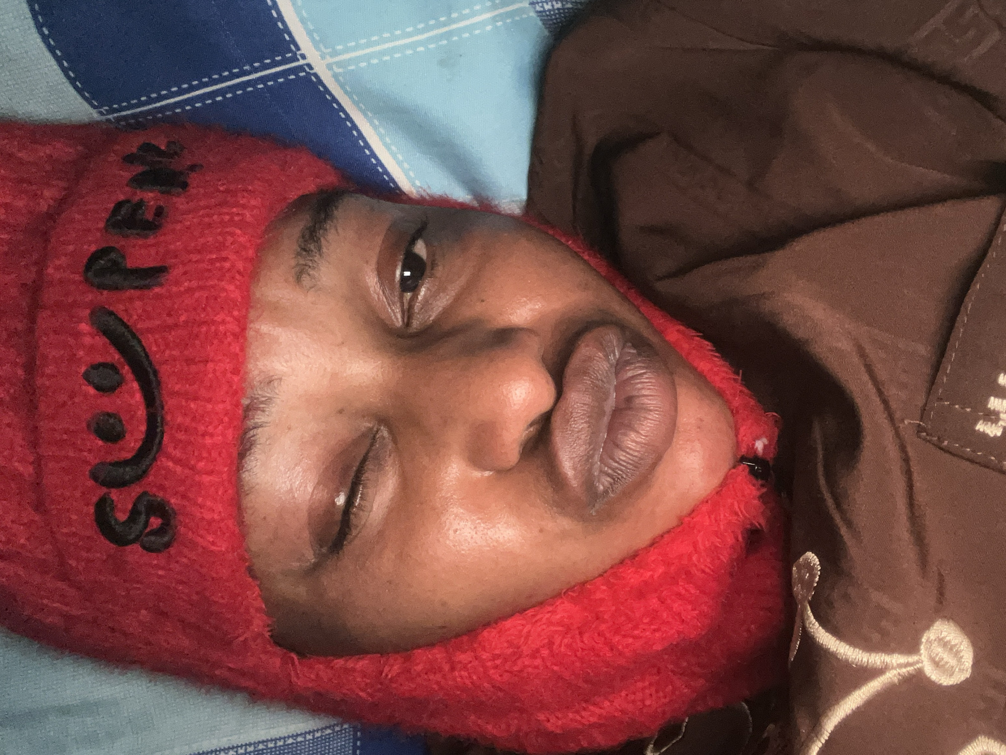 A close-up, over-the-shoulder selfie shows a person wearing a red balaclava with the word "SUPER" embroidered on it in black lettering. The person's face is partially obscured by the balaclava, with only their eyes and mouth visible. One eye is closed, and the other is open and looking directly at the camera. Their lips are pursed. The person is lying on a blue and white checkered bed. The scene is set in Jos, Nigeria, as indicated by the presence of a traditional embroidered brown garment worn by the subject. The lighting is dim, suggesting it might be nighttime or an indoor setting with minimal illumination.