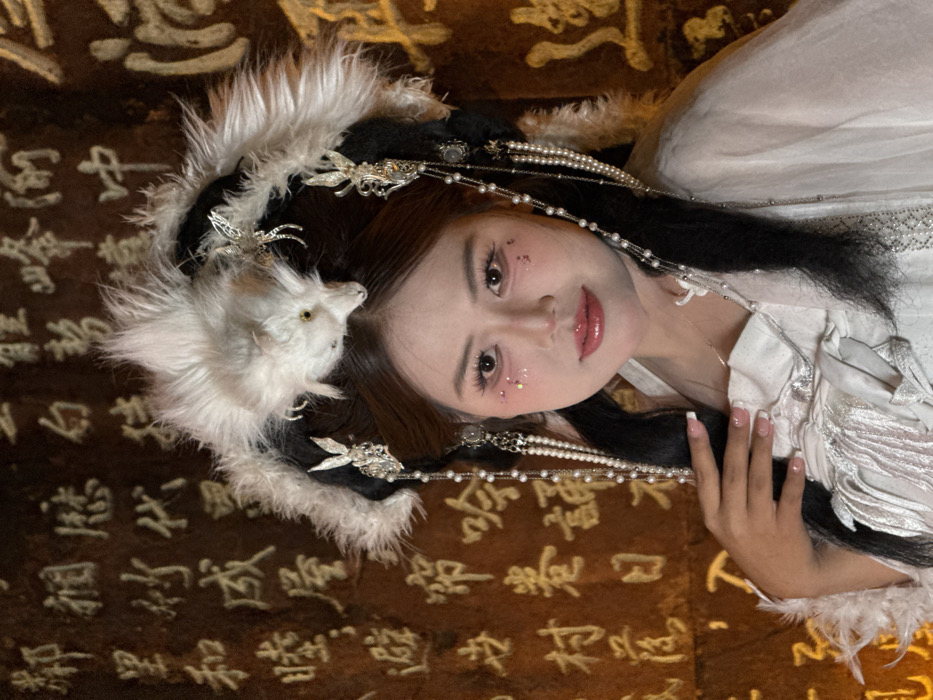 A female individual, appearing to be of East Asian ethnicity, is centrally positioned in a medium shot, facing the viewer with a slight smile. Her dark hair is elaborately styled with two side buns, a top knot, and a prominent white and black faux-fur headpiece resembling a stylized animal, possibly a fox, featuring small yellow eyes. Silver-colored decorative hairpins, some depicting rabbits, and multiple strands of white pearls and beads cascade from the headpiece. She wears a multi-layered, predominantly white garment, indicative of traditional Chinese attire, with textured fabric details. Her makeup includes pink eyeshadow, glitter on the upper cheeks, and glossy pink lips. Her right hand, exhibiting French-manicured nails, is gently placed near her left shoulder. The background is a dark, reddish-brown stone wall intricately carved with illuminated Chinese characters, consistent with a cultural or historical site in Yuzhong District, China. The scene is illuminated by warm, artificial light.