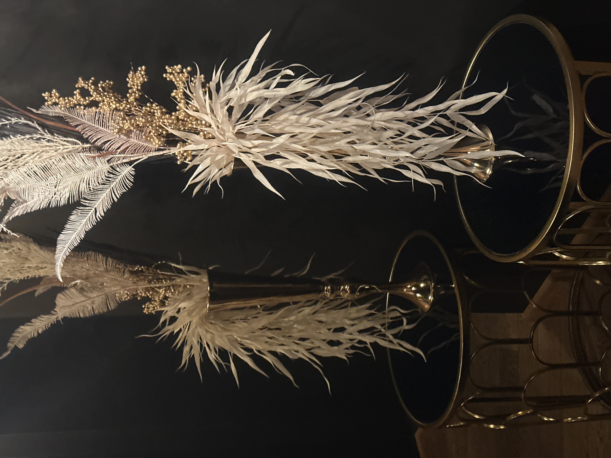 The image shows a floral arrangement with white and gold elements in a trumpet-shaped vase. The vase is placed on a black, reflective surface, creating a mirrored image of the arrangement and the vase. A gold-colored metal table with a circular top is partially visible on the right side of the image, also reflected in the surface. The background is dark. There is no evidence to suggest the location is Riga, Latvia.