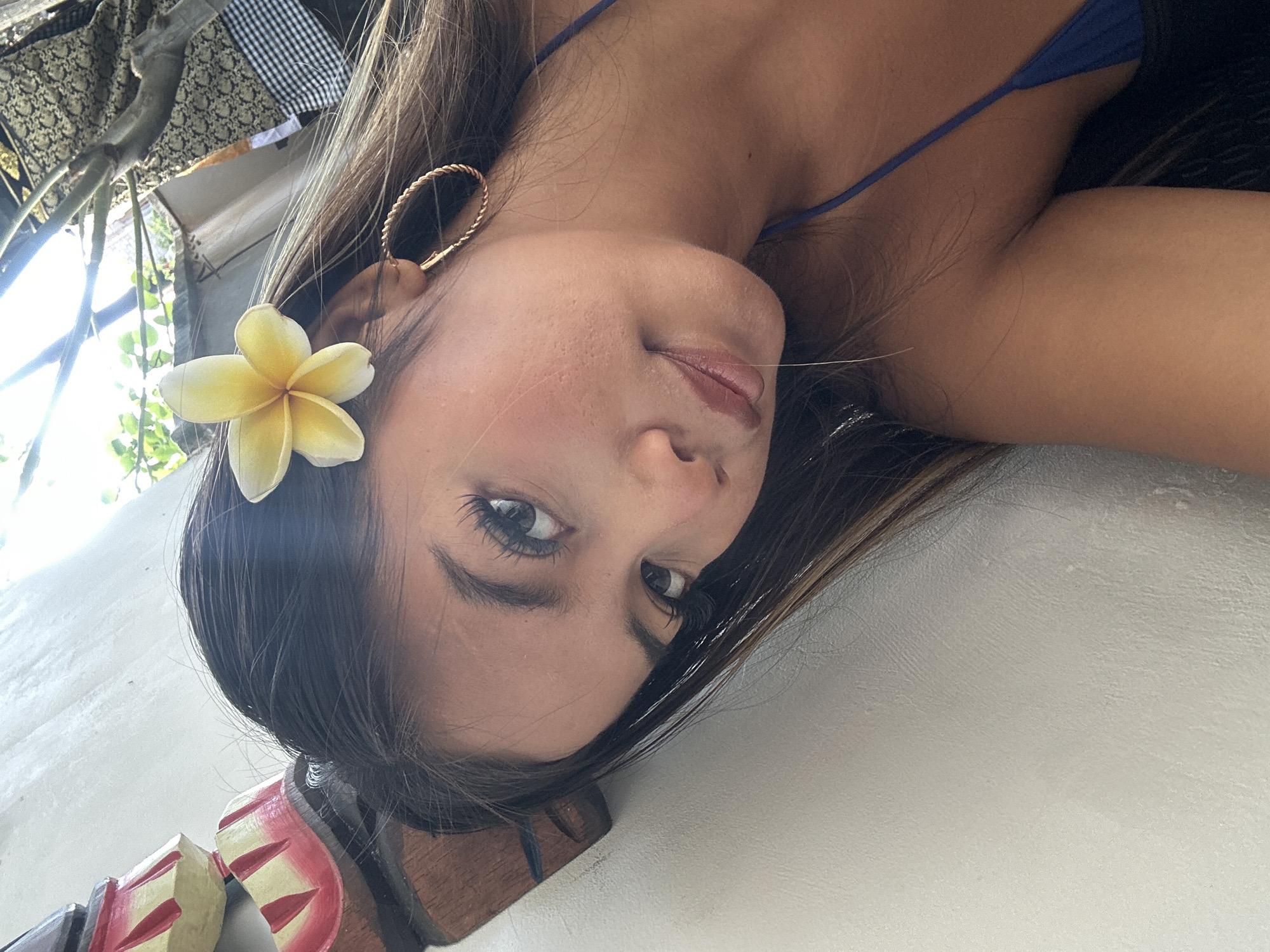 The image is a close-up, outdoor portrait of a young woman in Sukawati, Indonesia. She is positioned leaning against a textured, light-colored wall, with a blurred background that suggests an outdoor setting with foliage and some patterned fabric. The woman has dark hair, a frangipani flower tucked behind her right ear, and is wearing a blue bikini top. Her gaze is directed towards the camera with her head tilted. She has dark eyeliner and mascara, and her lips are slightly parted. A gold hoop earring is visible in her right ear. In the upper left portion of the frame, a decorative wooden object with red and yellow painted elements is partially visible, leaning against the wall. The lighting suggests it is daytime, with sunlight filtering in from the upper right.