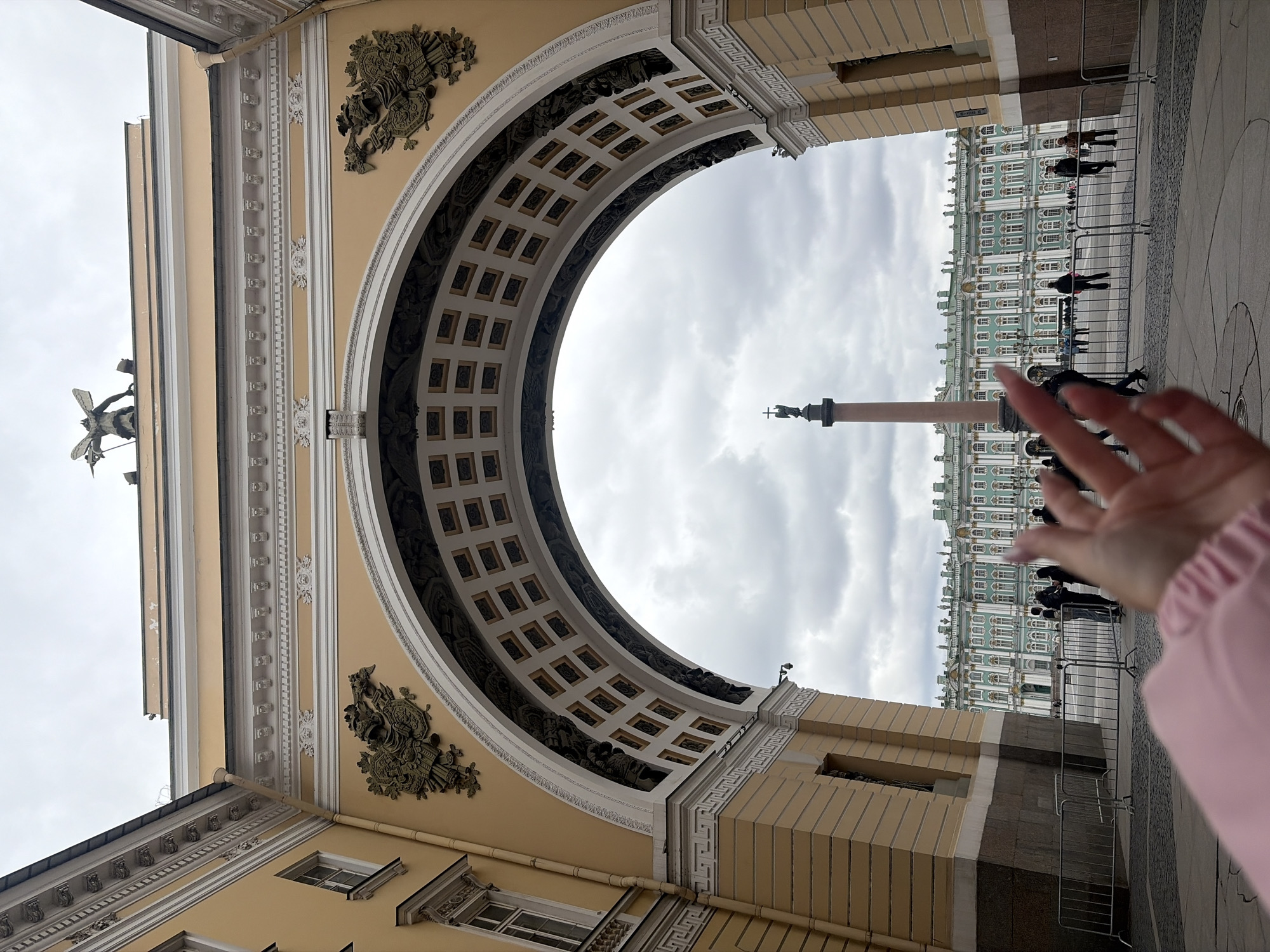 The image, presented in a vertical orientation, frames a monumental archway of a grand building from a low perspective, looking out towards a historic square. The archway, part of the General Staff Building in Saint Petersburg, Russia, is richly decorated with classical architectural elements, including a coffered soffit, intricate moldings, and ornate relief sculptures on its spandrels. The structure is painted in a light yellow hue with contrasting white details.
In the immediate foreground on the right side, a human hand, wearing a pink sleeve, reaches into the frame. Beyond the archway, the iconic green and white facade of the Winter Palace (Hermitage Museum) stretches across the background. To the left of the palace, the tall Alexander Column, topped with a statue, is visible against the sky. Several indistinct figures of people are seen walking on the paved ground of the square, with some separated by metal crowd control barriers. On the upper left of the arch's adjacent structure, a statue of a winged figure stands against the sky.
The scene takes place during the daytime under an overcast sky, casting a soft, diffuse light over the historical architecture. The prominent landmarks clearly identify the location as Palace Square in Saint Petersburg. There is no clearly legible text visible in the image.