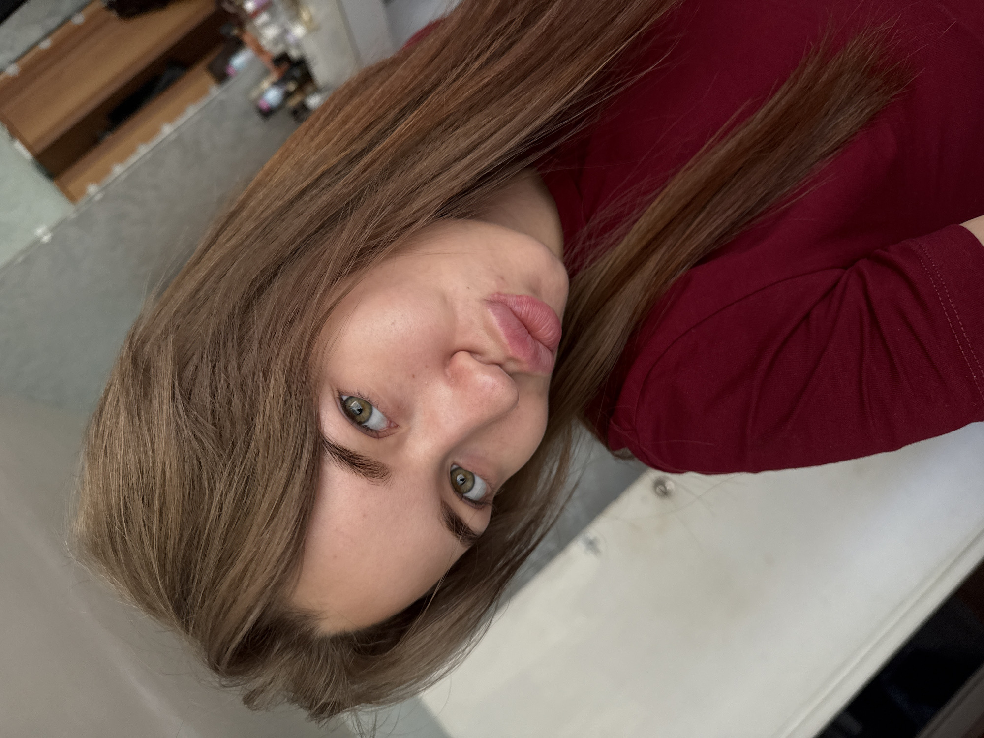 The image is a close-up selfie of a young woman with long, light brown hair and green eyes. She is wearing a dark red shirt and has her lips puckered. The background appears to be an indoor setting, possibly a bathroom or dressing area, with a mirror visible in the upper right corner reflecting a wooden cabinet and bottles of toiletries. The lighting is soft, and the overall impression is casual and personal. The image does not provide any specific details or visual cues that would allow for identification of the location within the city of Chaikovsky, Russia.