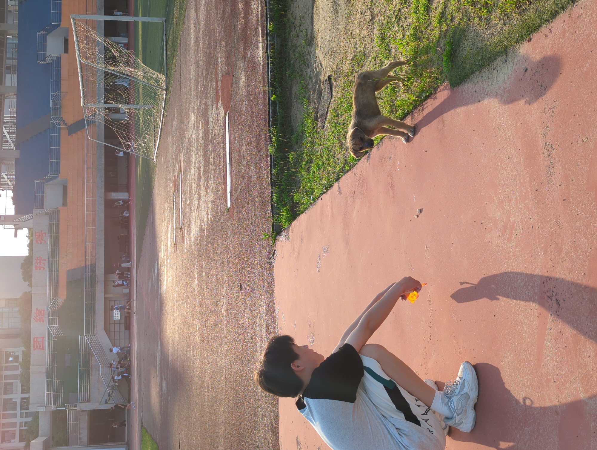 A low-angle, long-shot photograph taken outdoors at what appears to be a school or sports complex in Jiangjin, China, shows a young person, a dog, and a goal post on a red-colored track. The young person is seated, facing away from the camera, and is holding an orange object in their hands, possibly blowing bubbles. The dog is standing on a grassy area adjacent to the track. A goal post is visible further away, near a multi-story building with blue, orange, and white sections. The scene is bathed in sunlight.