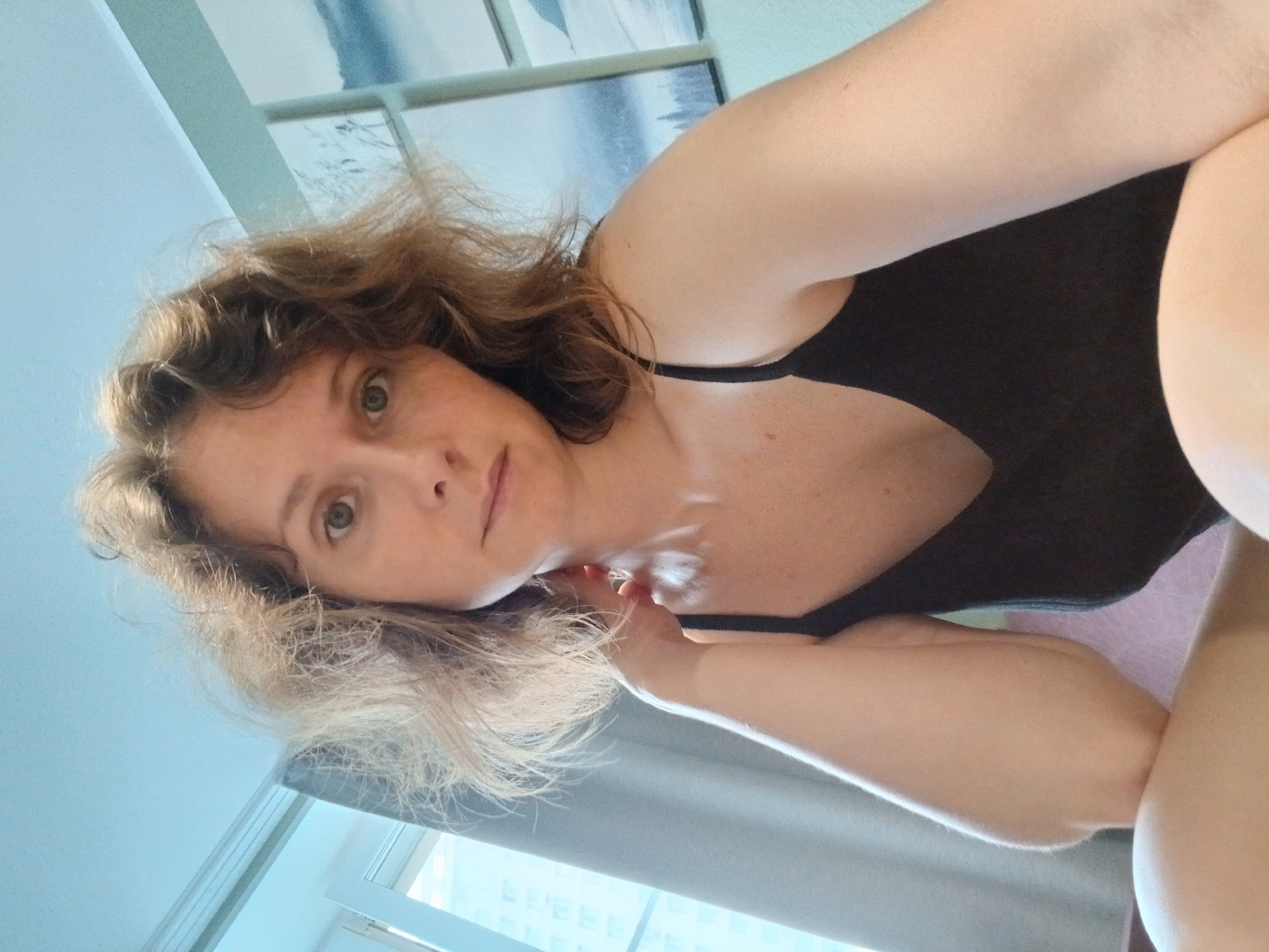 A woman with light brown, wavy hair and green eyes is shown in a close-up selfie. She is wearing a black tank top and her left arm is bent, with her hand resting on her neck. The background includes a window with a sheer gray curtain, a light blue ceiling, and two framed pictures on the wall depicting snowy landscapes. The lighting suggests it is daytime, with natural light coming from the window. The scene appears to be indoors, possibly in a residential setting. There is no information to suggest this image was taken in Yekaterinburg, Russia.