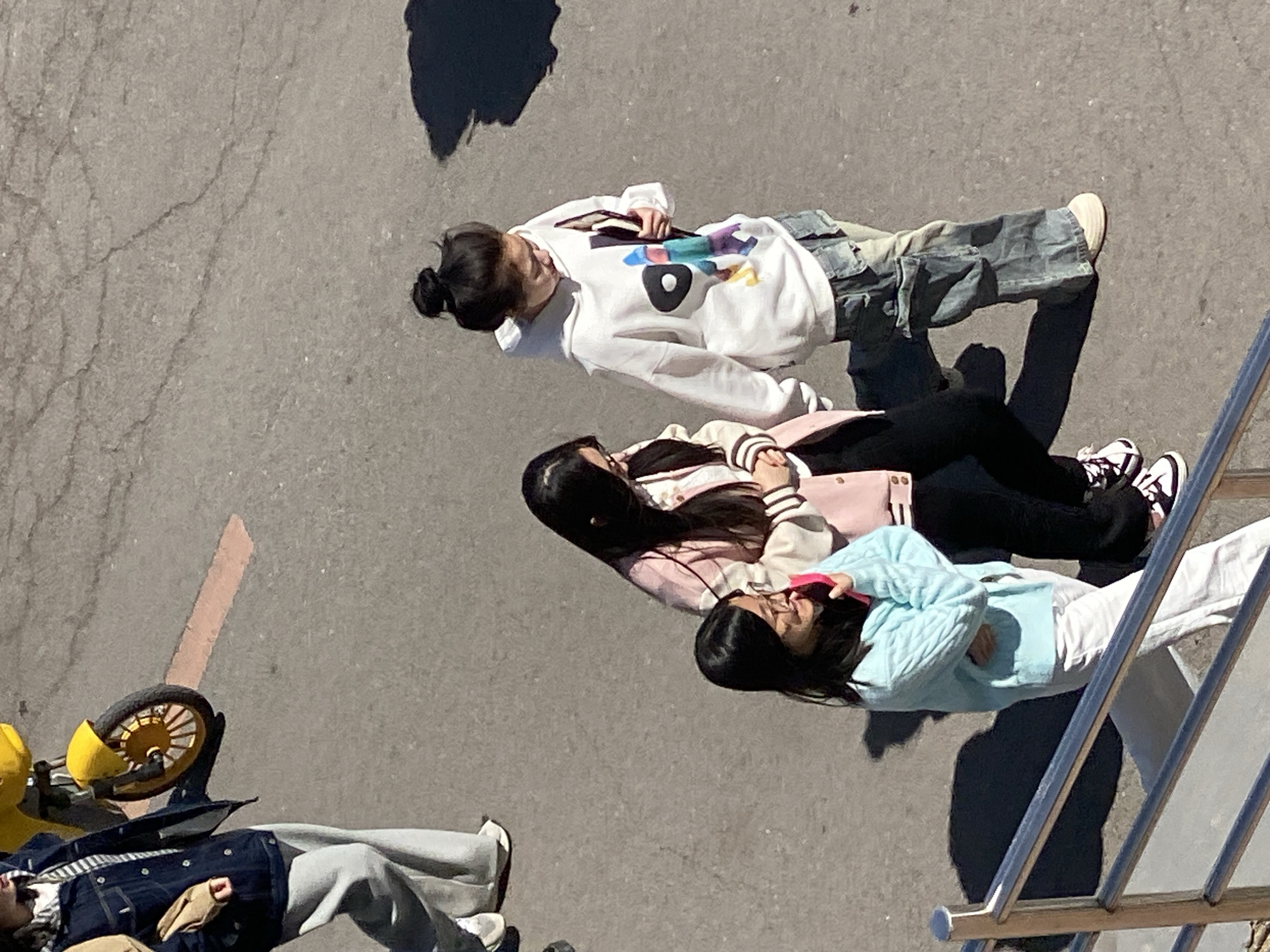 The image shows a view from above of a group of four young women lying on a paved surface, possibly a sidewalk, in the city of Zhongtun, China. The weather is bright and sunny.
The first woman, at the top of the frame, is wearing a white sweatshirt with colorful graphics, cargo pants, and sneakers. Her hair is in a bun. She appears to be holding a book or phone.
The second woman is wearing a pink and white varsity jacket, black pants, and black and white sneakers. Her long black hair is loose.
The third woman is wearing a light blue sweater and white pants. Her long black hair is loose, and she appears to be resting with her arms crossed.
The fourth woman, at the bottom of the frame, is wearing a blue jacket, a striped shirt, and gray pants. She is positioned near a yellow child's scooter.
A metal railing is visible in the right side of the frame. There is a dark shadow above the first woman. A reddish marking or line is visible to the left of the fourth woman. The overall environment appears to be urban, with a gray paved surface being the prominent feature.