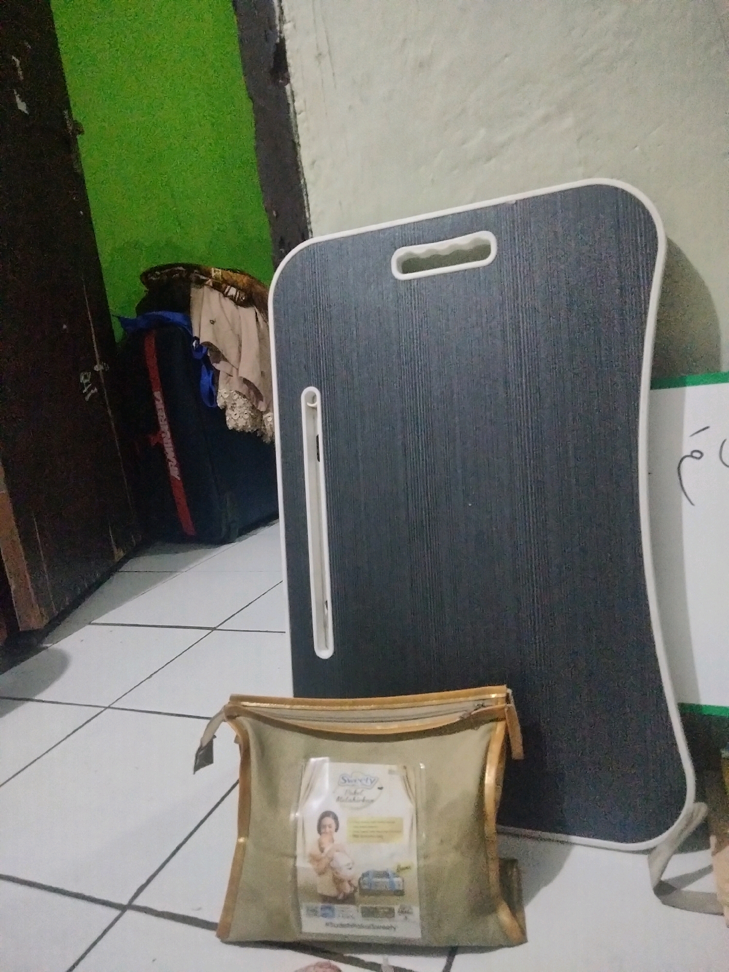 The image depicts an indoor scene, possibly a room, with tiled flooring. In the foreground, there is a light-colored zippered pouch with brown trim. On the pouch, a label features an illustration of a woman holding a baby. Behind the pouch, a dark gray lap desk with a white trim and a white handle is propped against a white board with some writing on it. To the left of the lap desk, there is a large black bag with red and white accents, and some clothing is draped over it. The wall behind these items is a vibrant green. The overall lighting suggests an indoor environment, and the presence of a lap desk and bag might indicate a personal space or storage area. There are no people clearly visible in the image. The location is stated to be Suka Sari, Indonesia.