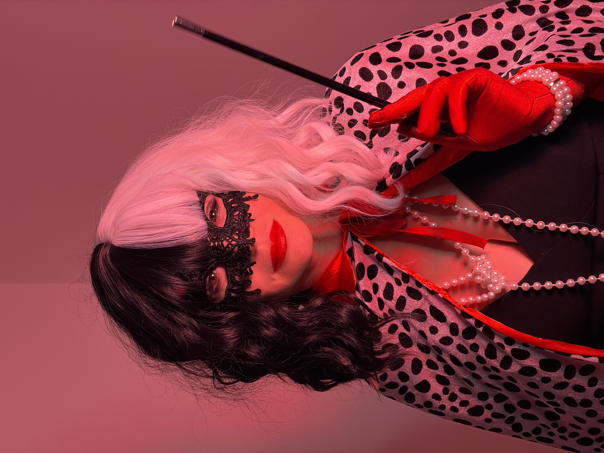 A woman dressed as Cruella de Vil poses against a red background. She wears a black-and-white wig, a black lace mask, red lipstick, a black dress, a pearl necklace, a Dalmatian-print shawl with red trim, and red gloves with a pearl bracelet. She holds a long black cigarette holder in her gloved hand. There is no indication of the city of Budva, Montenegro, or any other location within the image.