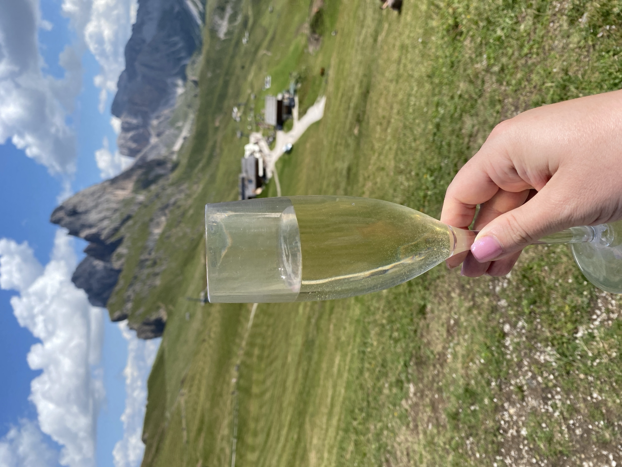 In Santa Cristina Gherdëina - St. Christina in Gröden - Santa Cristina Valgardena, Italy, a hand with bright pink fingernails holds a champagne flute filled with a pale yellow, bubbly liquid. The glass is positioned in the foreground, obscuring a portion of the backdrop. The background reveals a lush, green mountainous landscape under a partly cloudy blue sky. Jagged, rocky peaks dominate the distant horizon, and a cluster of small buildings, likely chalets or huts, are visible on the mountainside, with a road winding between them. The overall scene depicts a serene and picturesque mountain setting.