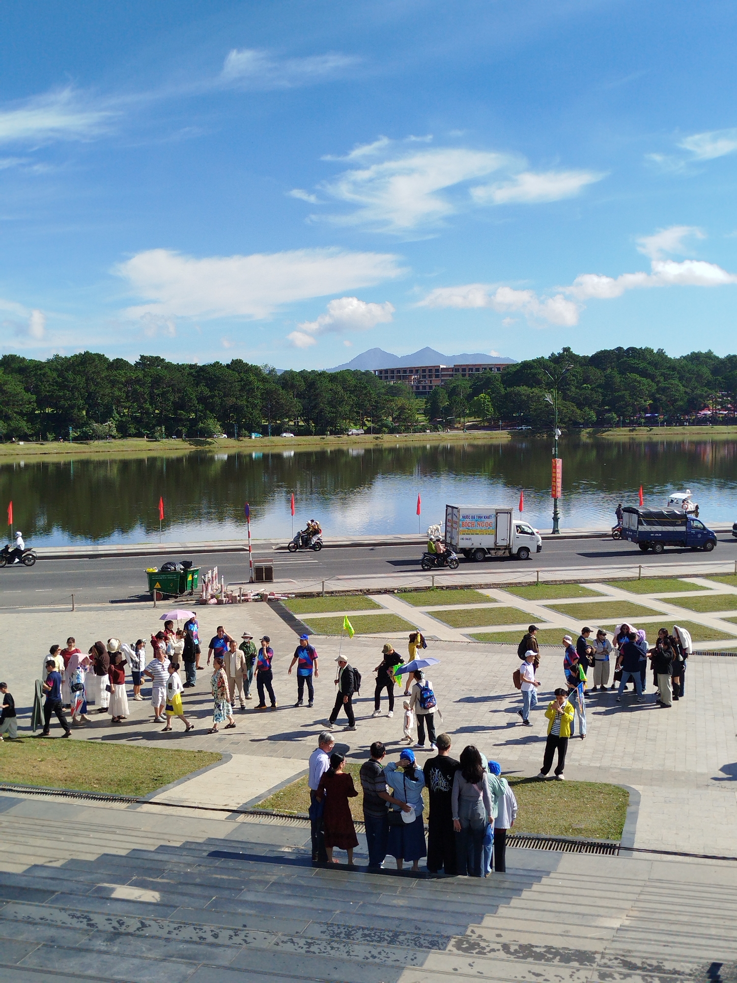 The image depicts a wide, scenic view of Xuan Huong Lake in Da Lat, Vietnam, on a bright, clear day with scattered white clouds. In the foreground, a group of people are gathered on a wide stone staircase, some looking towards the lake and others interacting amongst themselves. The mid-ground shows a road bordering the lake, with several vehicles including motorcycles, a white truck, and a blue truck. People are seen walking and riding on the motorcycles. The lake itself is calm and reflects the surrounding trees and sky. Beyond the lake, a dense line of pine trees forms a lush green backdrop, with buildings visible in the distance, hinting at the urban landscape of Da Lat. The overall atmosphere is one of a public gathering space, likely a tourist area, with a mix of locals and visitors enjoying the outdoor environment.