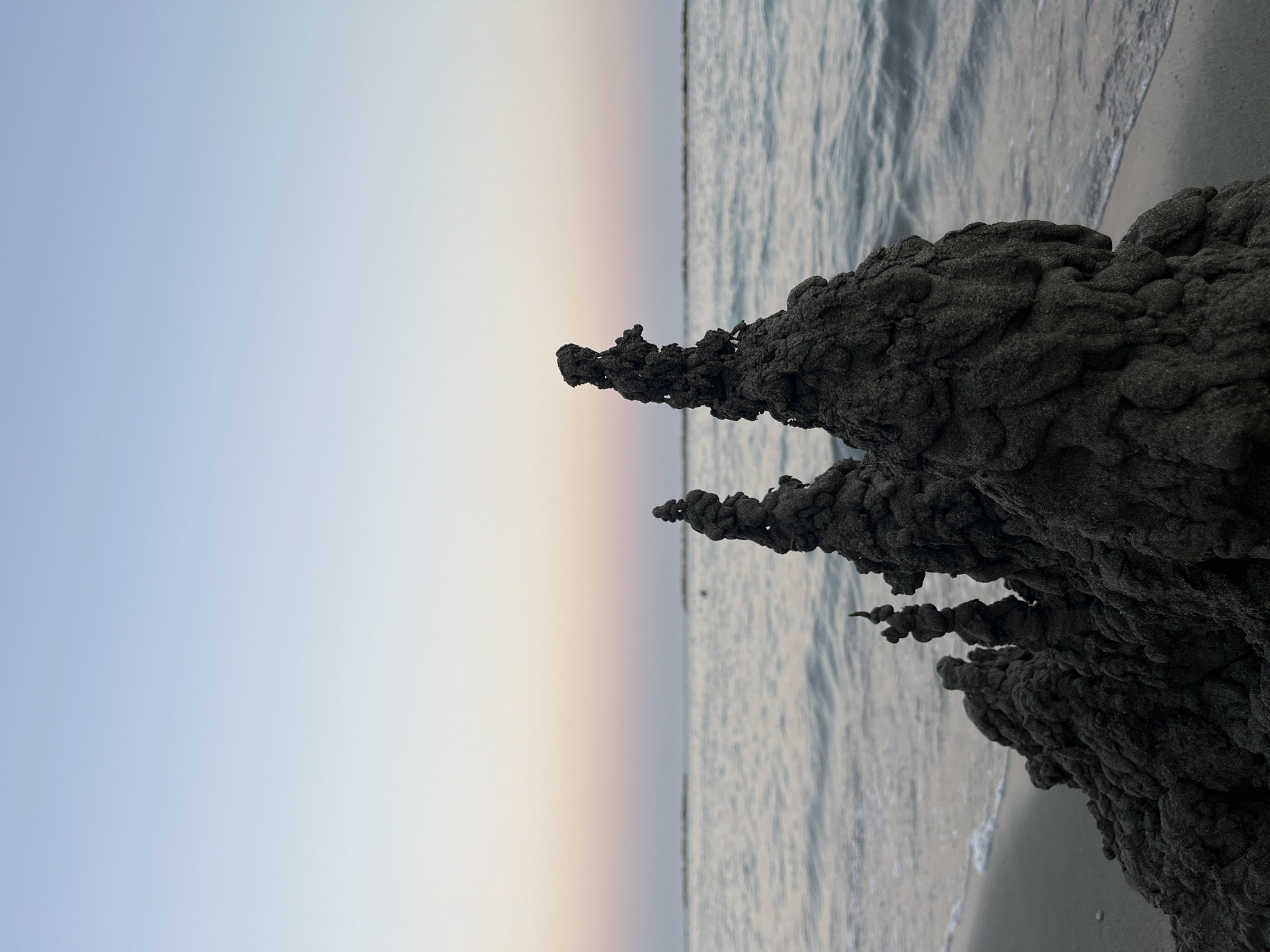 The image depicts a cluster of dark, intricately formed sand structures on a beach, likely at sunset, given the soft gradient of pastel colors in the sky and the calm water. The sand structures resemble miniature, abstract towers or spires, with a rough, textured surface. In the background, the sea meets the horizon, with subtle waves and a hint of a shoreline. The sky transitions from a pale blue at the top to soft pink and peach hues closer to the horizon. The scene is tranquil and natural, evoking a sense of peace. The specific location of Agios Tychona Community, Cyprus, is not discernible from the visual information presented in the image.