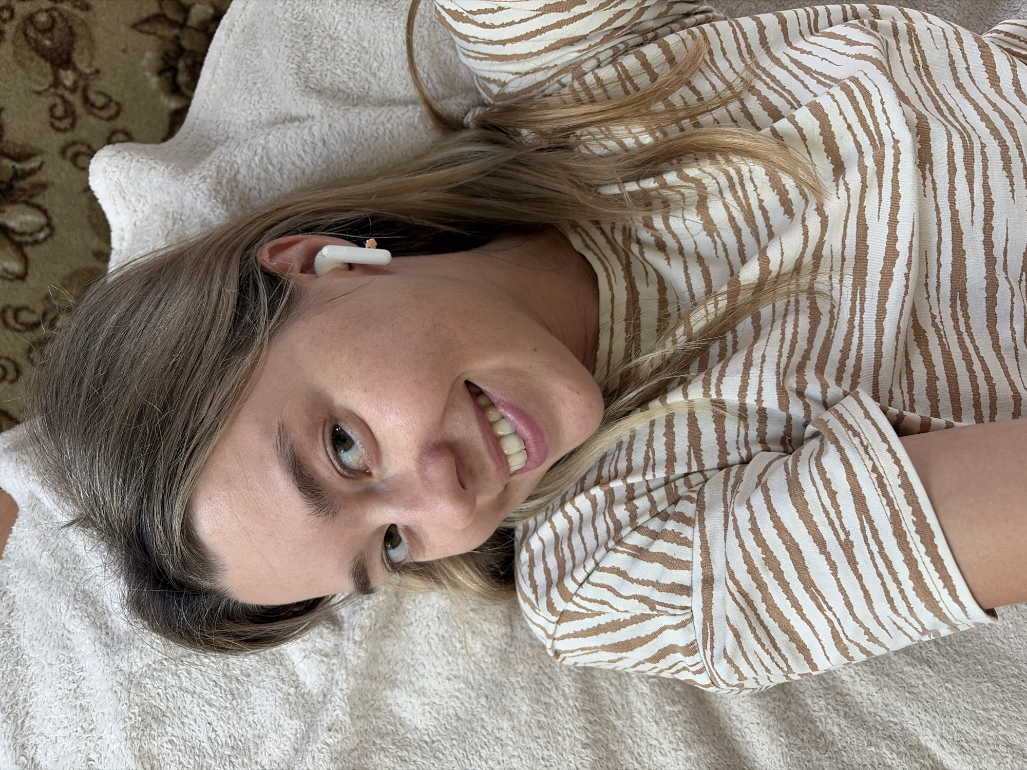 A young Caucasian woman with blonde hair is lying on her back on a white, textured blanket. She is wearing a brown and white striped t-shirt with a zebra-like pattern. The woman has a white wireless earbud in her right ear. Her head is slightly tilted to her left, and she is smiling directly at the camera. The background is a blurry patterned carpet. The scene appears to be indoors, possibly a home environment. The lighting is bright and natural, coming from above and slightly to her right. There is no indication of the city of Chaikovsky, Russia in the image itself.