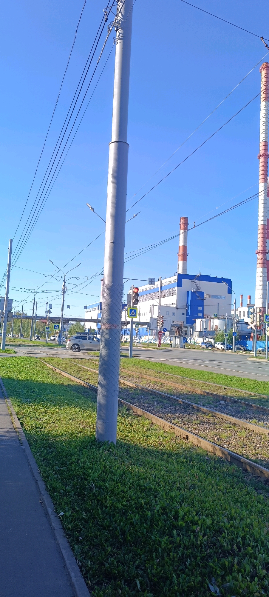 This is a medium shot, taken outdoors on a clear, sunny day. The scene depicts an urban environment in Perm, Russia, likely near an industrial area, as evidenced by the large factory building and industrial chimneys in the background.
In the foreground, a concrete sidewalk runs along the left edge of the image. Adjacent to the sidewalk is a patch of green grass, which transitions into a set of tram tracks. A large, grey utility pole stands prominently in the middle of the frame, with multiple electrical wires extending from it across the sky.
In the mid-ground, there is a road with several vehicles visible. A light-colored SUV is parked on the road, and other cars are moving in the distance. Traffic lights are visible, one of which is illuminated red. Pedestrian crossing signs are also present.
The background features a large, modern industrial building, painted in white and blue. Two tall industrial chimneys, striped red and white, rise above the building, indicating it is likely a power plant or similar facility. The sky is a clear, bright blue. The overall impression is of a functional urban landscape during daylight hours.