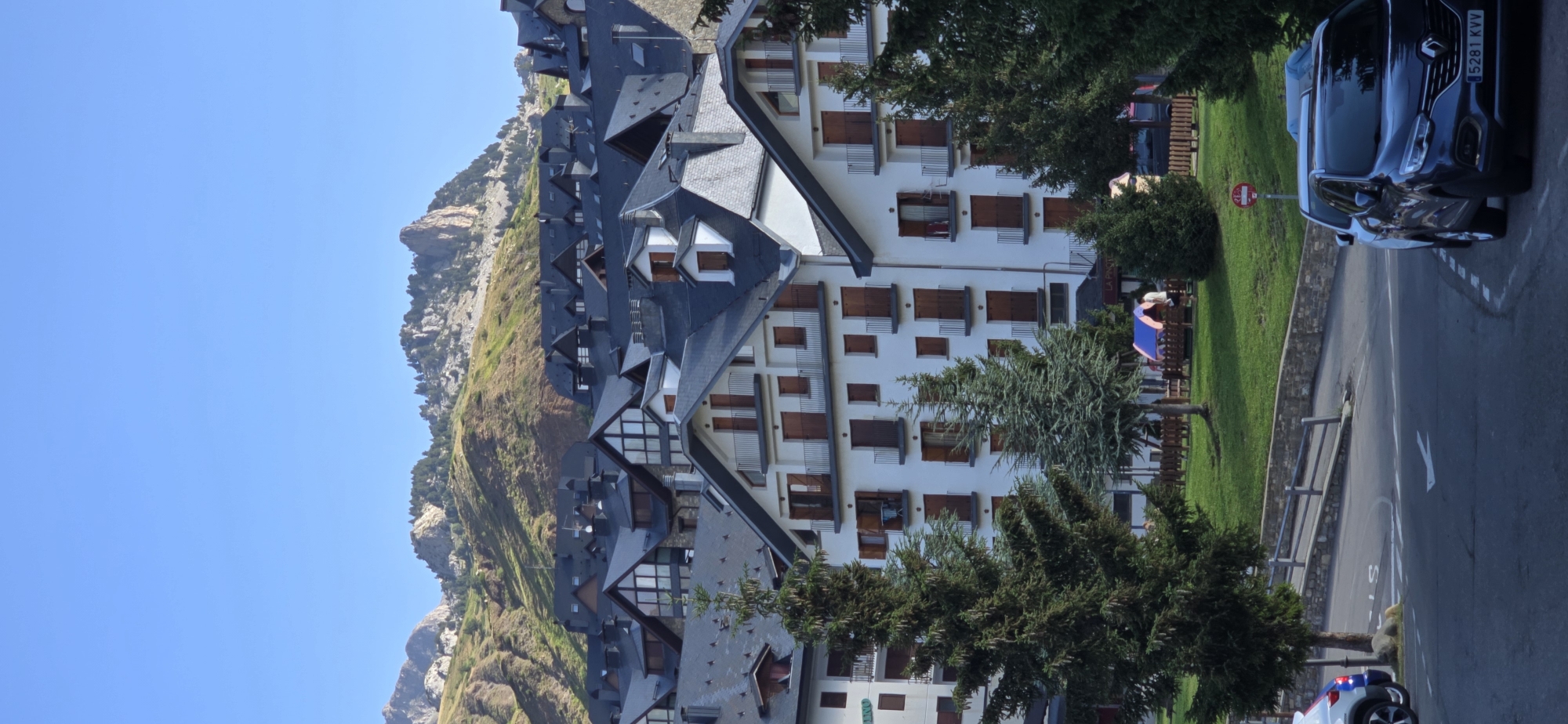 The image depicts a sunlit daytime scene in Sallent de Gállego, Spain, featuring a cluster of multi-story buildings with dark, angled roofs, characteristic of mountain architecture. The buildings are white-washed with visible balconies and dark-framed windows and shutters. In the foreground, two evergreen trees stand prominently, with lush green grass and a low stone wall separating the buildings from a paved area where several cars are parked. The mountainous terrain behind the buildings is covered in scrub and trees, leading up to rugged, rocky peaks under a clear blue sky. A red and white sign is visible near the parking area.