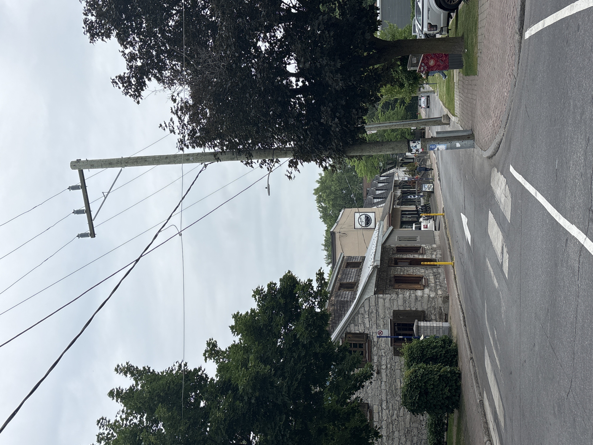 This is a street scene in Pointe-Claire, Canada, featuring a stone building with wooden shutters on the left, a paved road with a crosswalk, and a sidewalk with a red recycling bin on the right. Overhead, power lines are supported by a utility pole. In the background, there are several storefronts, trees, and parked vehicles. The sky is overcast. A person is visible near the recycling bin, and a couple of figures are walking on the sidewalk in front of the shops.