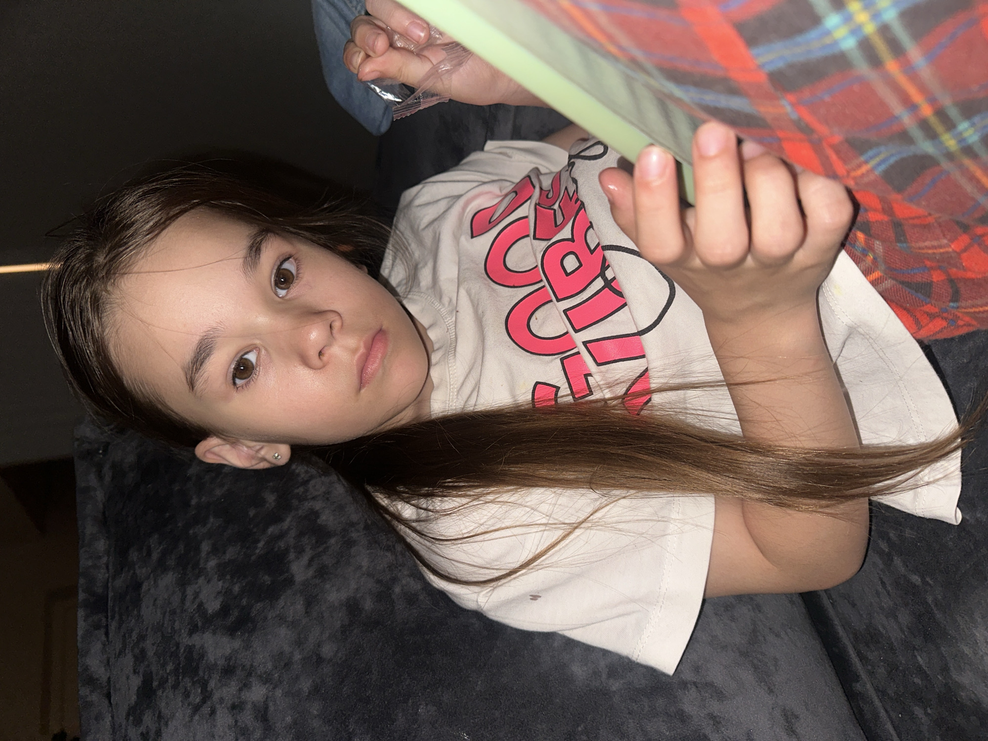 A young girl with brown hair and brown eyes is lying on a gray upholstered surface, possibly a couch or a bed. She is wearing a white t-shirt with a pink graphic design. She is holding a rectangular object with a green frame. Part of a plaid blanket is visible. The environment appears to be indoors, with dim lighting. The location of Kharnavy, Russia is not discernible from the image.