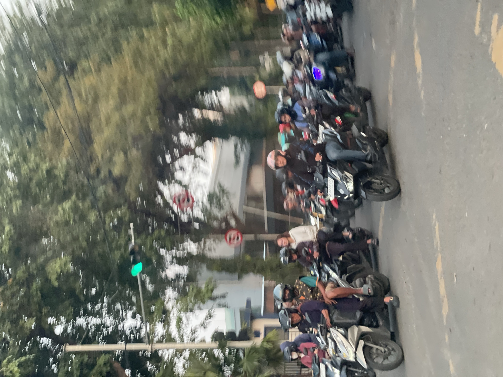 A blurry image, likely taken from a moving vehicle, shows a congested city street in North Jakarta, Indonesia. Several rows of motorcycles and scooters are parked along the roadside, with riders sitting on them. Many riders are wearing helmets. A traffic light displays a green signal. There are trees lining the street, with buildings visible in the background. A "no entry" traffic sign is visible in the background.