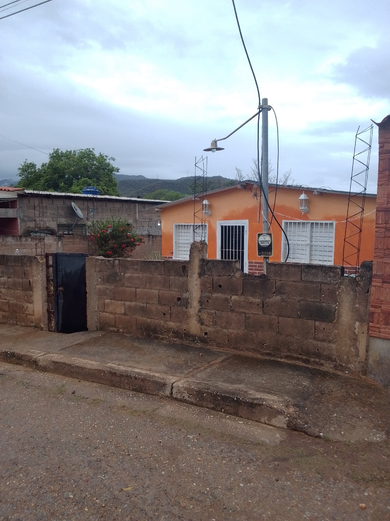 A wide shot captures a residential scene in Juan Griego, Venezuela, under an overcast sky, suggesting either early morning or a heavily clouded day. The foreground shows a rough, gravelly street with a concrete curb and a sloped driveway leading up to a brick wall. This wall, constructed from concrete blocks, partially obscures the view of a house and its yard. A dark, metal gate stands ajar within the wall on the left side.
Behind the wall, an orange-painted house with white-framed windows and doors is visible. The windows are barred for security, and two white outdoor light fixtures are mounted on the house's facade. A utility pole stands in front of the house, carrying electrical wires and a street lamp with a visible bulb. A utility meter is mounted on the pole at eye level. A cluster of red flowers blooms on a bush to the left of the house, contrasting with the muted tones of the surroundings.
Beyond the house, more buildings and structures can be seen, including a gray concrete building with a satellite dish on its roof and a metal structure resembling scaffolding or antenna supports. In the background, rolling hills and mountains are dimly visible through the hazy, cloudy atmosphere. The overall impression is of a somewhat modest, lived-in neighborhood under a somber sky. No people are visible in the frame, and no text is present.