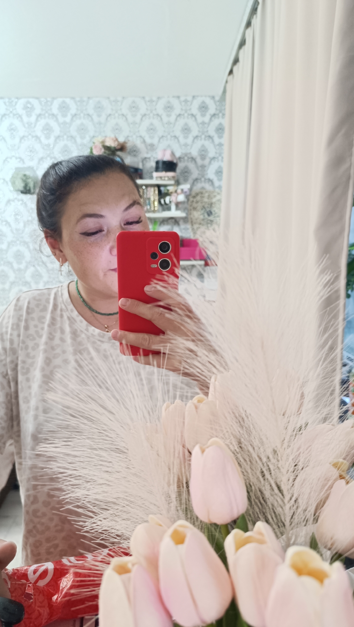 A woman with dark hair pulled back is taking a selfie in a mirror, holding a red smartphone. She is wearing a light-colored, leopard-print t-shirt and a green beaded necklace. She has freckles and her eyes are looking down at her phone. In the foreground, a bouquet of pale pink tulips and feathery plumes is visible. Behind her, reflected in the mirror, is a wall with a patterned wallpaper, shelves with decorative items, and a beige curtain. The scene appears to be indoors, possibly a dressing area or bedroom. There is no visible indication of the location being in Chaikovsky, Russia.