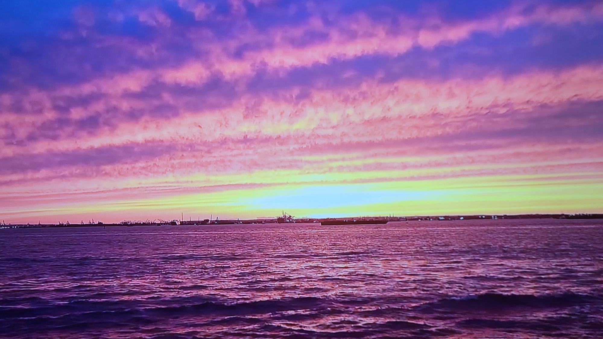 The image shows a sunset over water. The sky is filled with horizontal layers of clouds, ranging in color from purple and pink at the top to yellow and orange near the horizon. The water reflects the colors of the sky. On the horizon, there is a faint suggestion of land, with some industrial structures or buildings visible. The scene is not located in Yerevan, Armenia, as it lacks any recognizable features associated with that city.