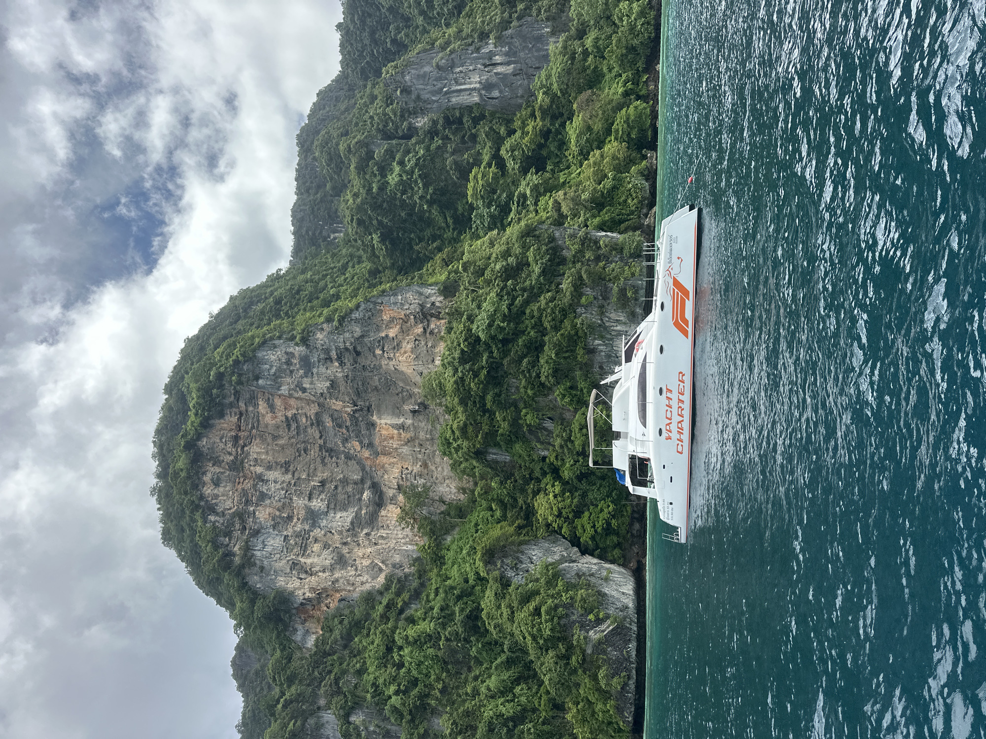 A white catamaran with orange lettering that reads "YACHT CHARTER" is docked in the emerald waters off the coast of Ao Nang, Thailand. The boat is positioned in the foreground, with its bow pointing to the left of the frame. In the background, a towering limestone cliff face covered in lush green vegetation rises dramatically from the sea. The sky above is overcast with scattered clouds.