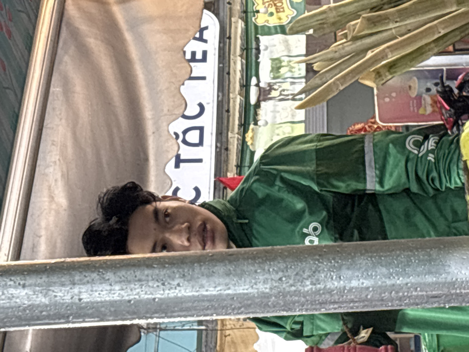 The image captures a young adult male in Ngũ Hành Sơn Ward, Vietnam, positioned behind a prominent, wet, cylindrical metal pole in the foreground. He is wearing a dark green, long-sleeved uniform jacket, identifiable by the white "Grab" logo on the left chest and reflective grey bands on the sleeves. His dark hair is styled, and he looks directly towards the viewer with a neutral expression and slightly parted lips. In the background, a white sign with a wavy top edge displays black text, partially legible as "TẮC TEA." Above the sign, a light-colored fabric awning provides overhead cover. To the right of the male subject, a display features multiple clear cups containing various beverages, alongside a bundle of cut sugarcane stalks. A green and yellow sign, possibly advertising "SỮA HỘT," is partially visible on the far right. A portion of a motor scooter's handlebar is discernible in the bottom right corner.