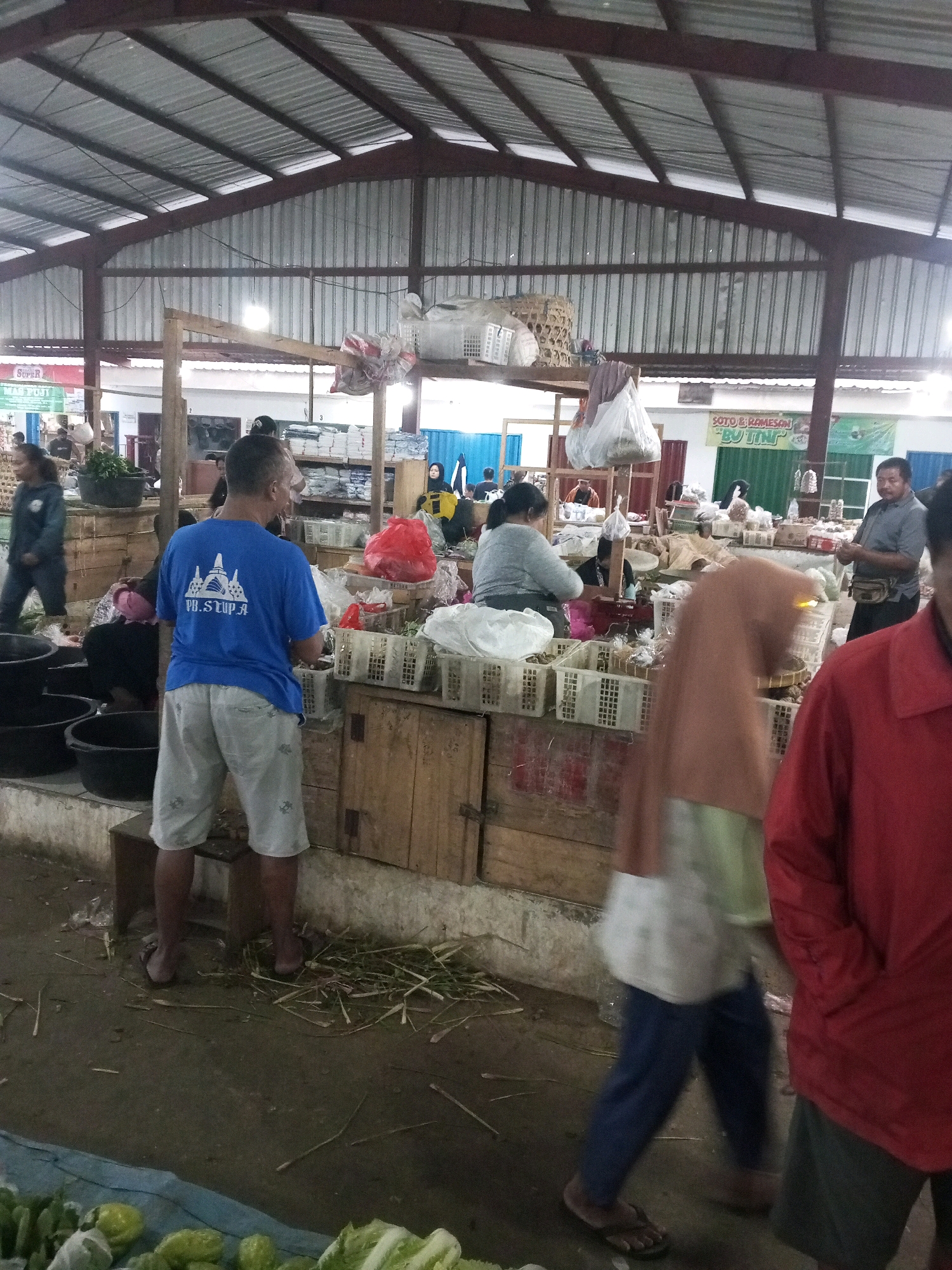 This image depicts an indoor market scene in Gentan, Indonesia. The market appears to be covered by a corrugated metal roof supported by metal beams and wooden structures. The stalls are arranged with wooden counters displaying various goods, primarily in plastic crates and baskets. Several individuals are visible, engaged in activities typical of a market.
In the foreground, a man wearing a blue t-shirt with a white logo and light-colored shorts is facing away from the camera, appearing to be a vendor or shopper. To his right, a woman in a light-colored hijab and patterned clothing is walking, her body blurred due to motion. Further to the right, a person in a red shirt is also partially visible, facing the side.
In the background, other market-goers and vendors are seen. A man in a gray shirt and dark pants with a fanny pack is standing near a stall. Another woman in a gray sweater is interacting with a stall. Various items are displayed, including what appears to be produce, packaged goods, and possibly clothing. Large black plastic basins are visible in the lower left corner. The ground is a dirt or concrete surface, with some debris and straw scattered around. Signs with text are present on the stalls and in the background, indicating the market's commercial nature.