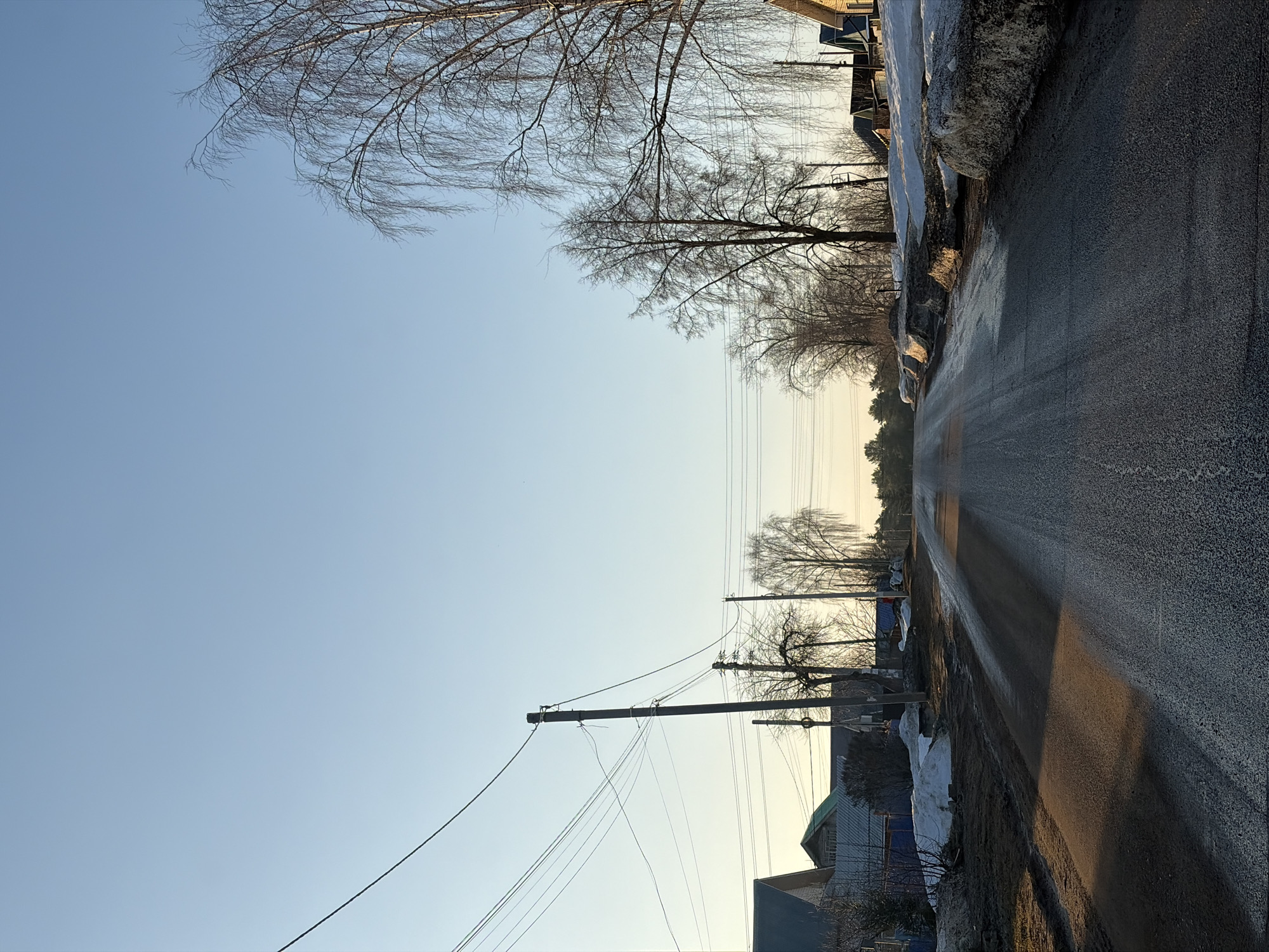 The image shows a street in Chaikovsky, Russia. The road is paved and runs straight into the distance, reflecting the sunlight. The sky is clear and pale blue. Trees line the street, their branches bare, indicating a winter or early spring setting. Power lines stretch along the road, supported by poles. Patches of snow remain on the sides of the road, next to houses.