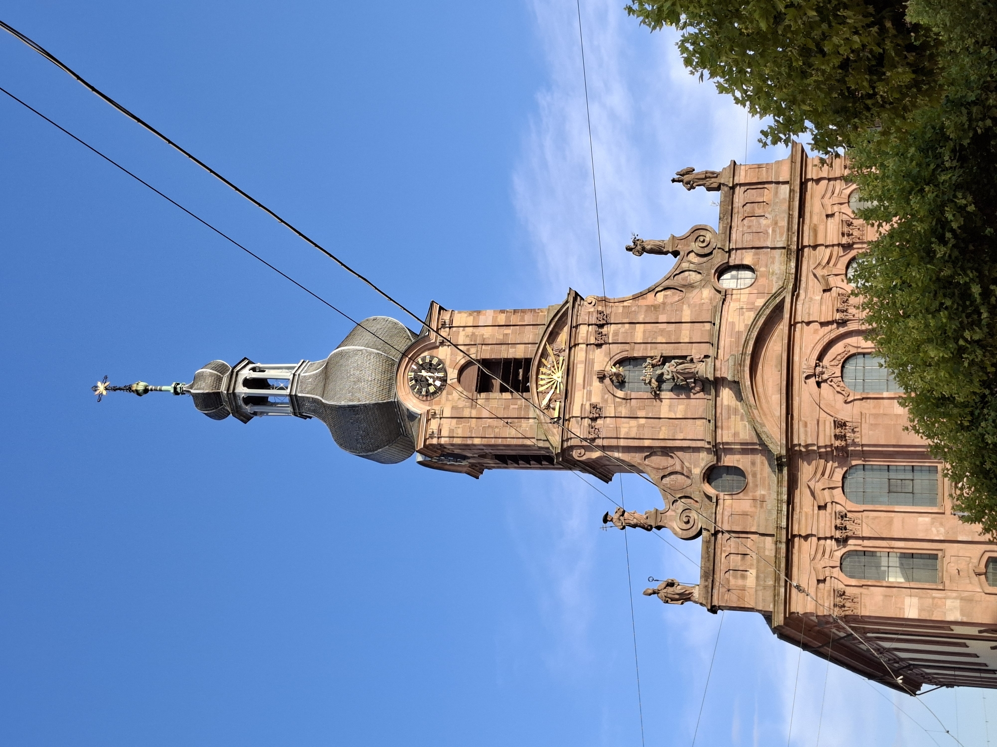 The image shows a close-up, low-angle view of the historic St. Alexander's Church in Rastatt, Germany. The church's prominent red sandstone facade is richly decorated with sculptures and architectural details. A tall tower with a clock and a domed spire topped with a cross rises against a clear blue sky with a few wispy clouds. Overhead, electrical wires are visible, crossing the frame diagonally. Lush green trees partially obscure the lower portion of the church. The overall impression is one of grandeur and historical significance.