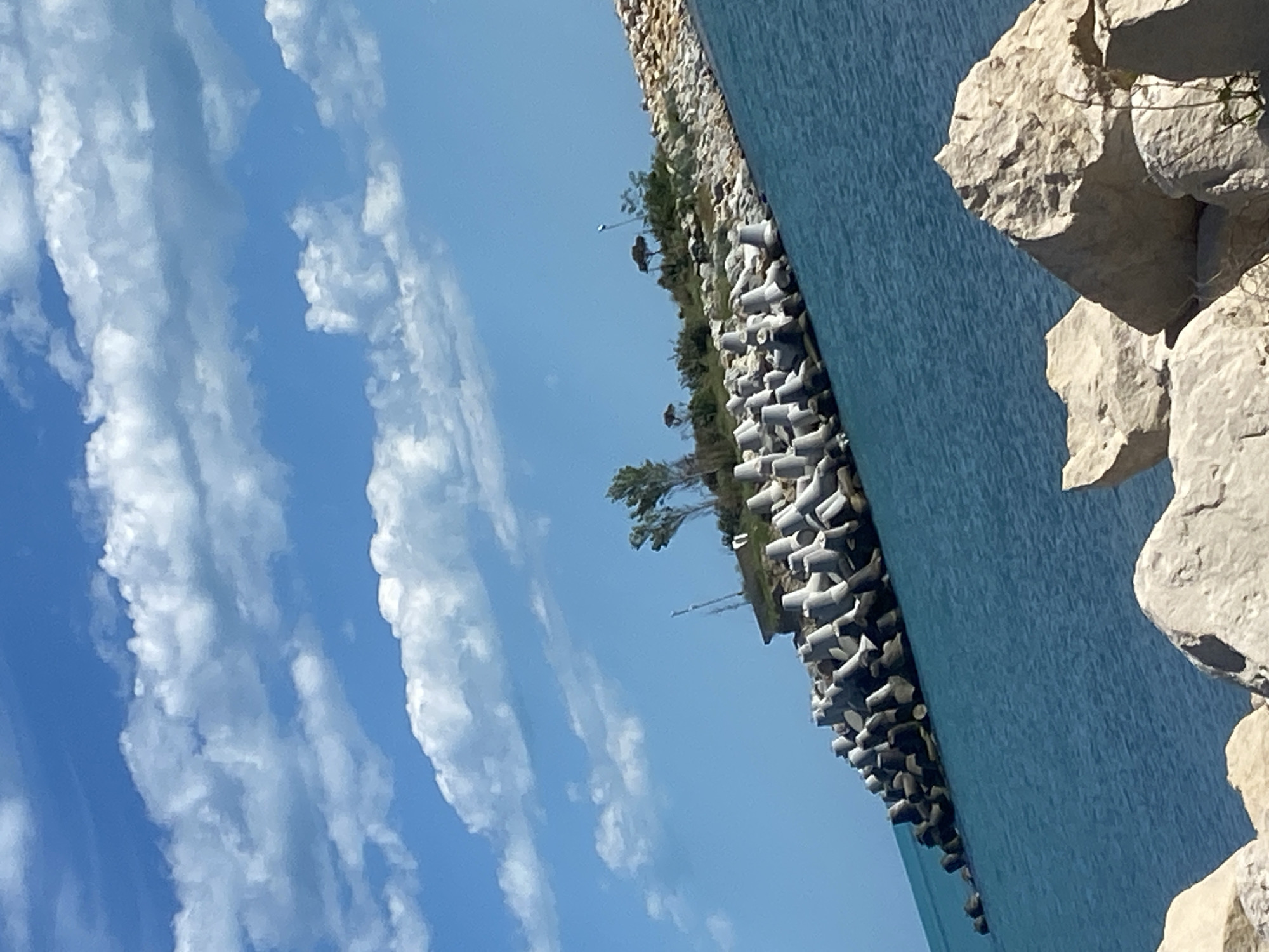 The image shows a coastline in Varna, Bulgaria, under a partially cloudy, blue sky. The right side of the frame features large, light-colored, roughly hewn rocks that form part of the shoreline or a breakwater. To the left of these rocks is a body of water, likely the Black Sea, appearing calm and slightly turquoise in color. Along the water's edge, there is a row of interlocking, geometrically shaped concrete structures, possibly tetrapods, designed to protect the coast from erosion. Behind these structures is a narrow strip of land with sparse greenery, including trees. In the background, utility poles can be seen. The viewpoint is tilted.