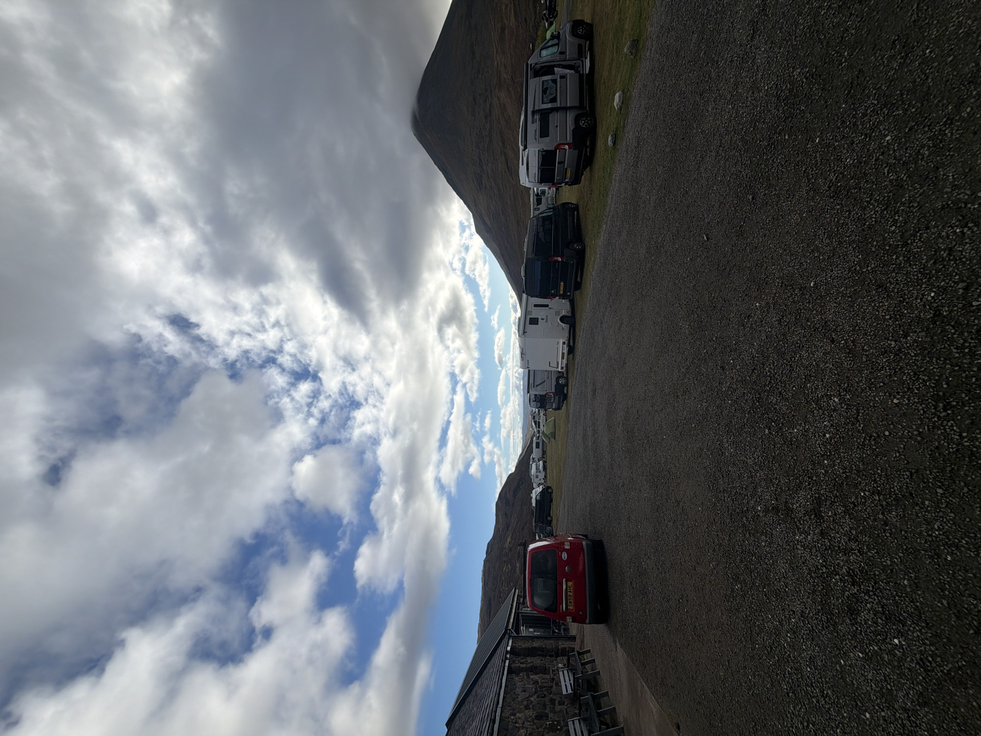 The image depicts a row of vans and campers parked along a gravel road in the Isle of Skye, United Kingdom. A red car is also parked nearby. The sky above features a mix of clouds and patches of blue. A mountain partially obscures the background. A building sits at the bottom edge of the image, next to the red car.