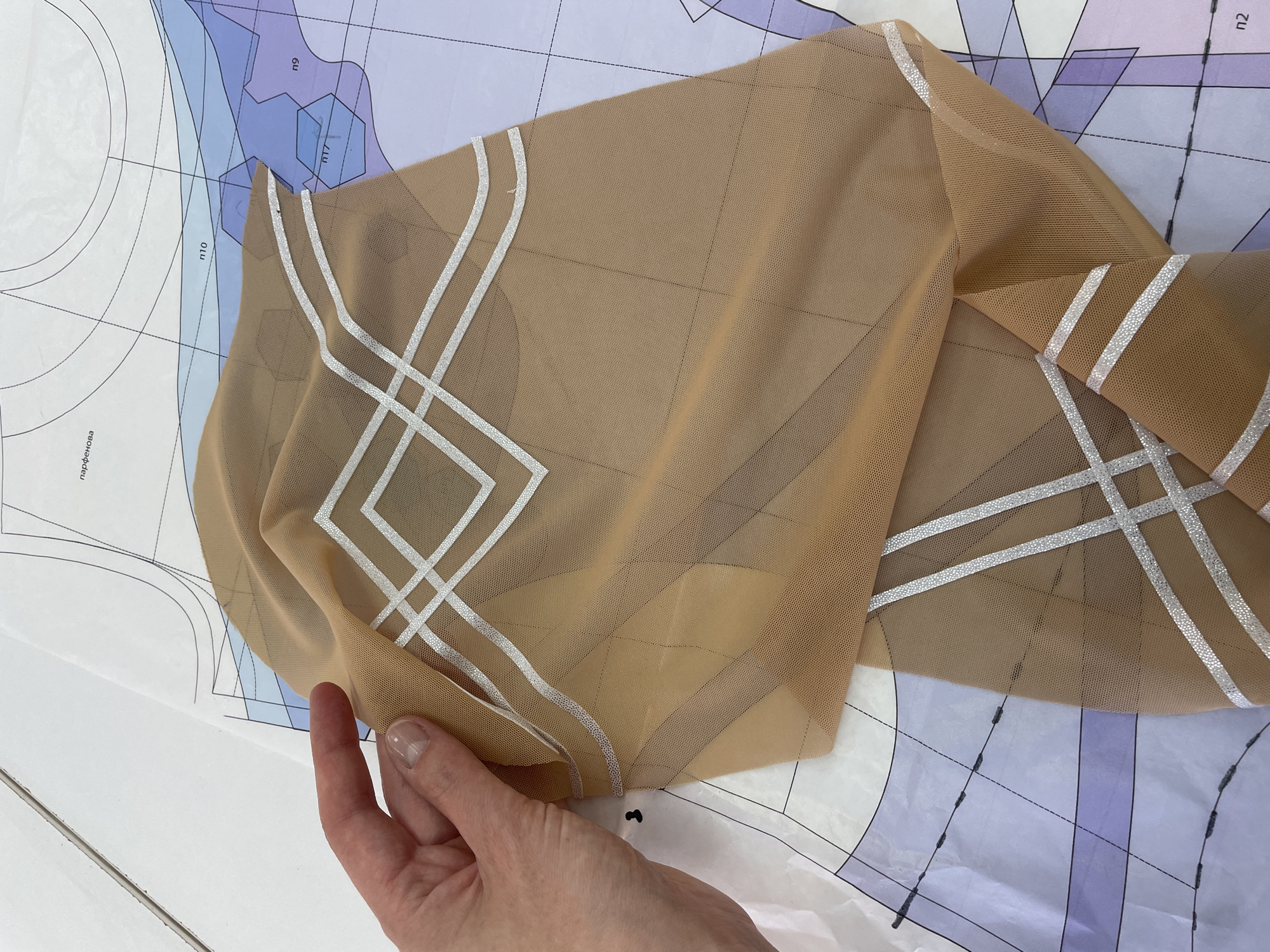 A hand holds a piece of sheer, nude-colored fabric with a geometric silver-foil pattern over sewing pattern paper. The fabric is draped and folded, obscuring much of the pattern paper beneath it. The pattern paper has various lines, color blocks, and numerical labels, suggesting it's a garment blueprint. The scene is photographed from a slightly overhead angle. The image does not provide any information about the location in Saint Petersburg, Russia.