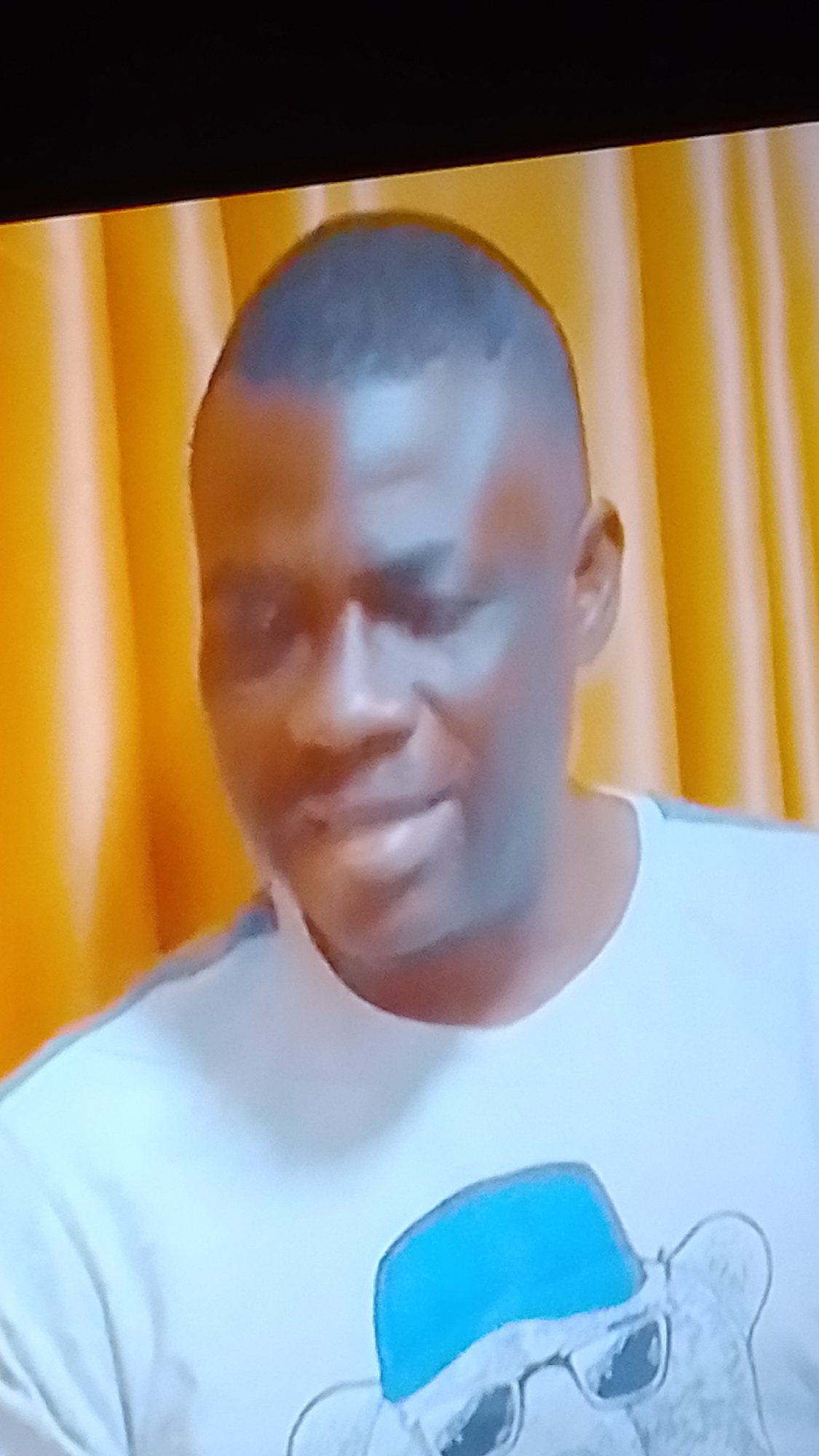 A close-up, waist-up shot shows a man of African descent with a closely cropped haircut and light stubble. He is wearing a light-colored t-shirt with a graphic of a bear wearing a blue cap and sunglasses. The background consists of vertical folds of yellow fabric, suggesting curtains or drapery. The lighting is uneven, with a bright light source to the left of the frame, casting shadows on the man's face. The image quality is grainy and appears to be captured from a screen.