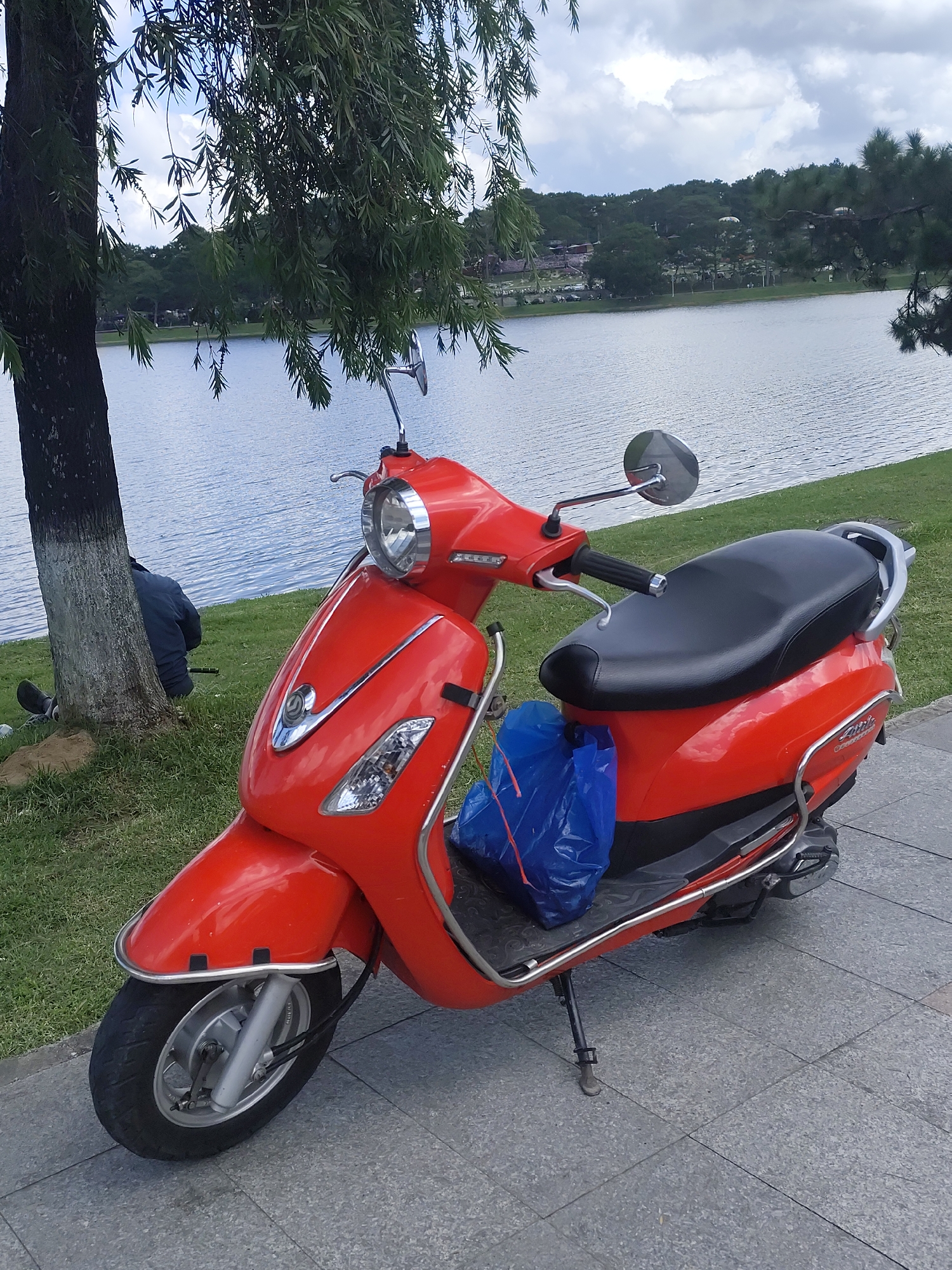 An orange scooter is parked on a tiled pathway next to a lake in Da Lat, Vietnam. The scooter has a black seat, a blue plastic bag on the rear rack, and chrome accents. A person in a dark jacket is sitting on the grass by a tree near the lake. The lake is calm and reflects the sky, which is partly cloudy. Trees and buildings are visible on the far side of the lake, suggesting an urban or park setting. The scene appears peaceful and recreational, with the scooter suggesting a mode of transport for exploring the area.