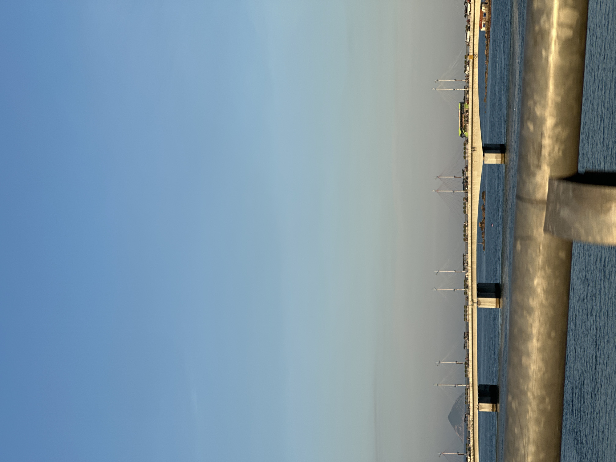 The image shows a view of the sea and a bridge, potentially in Bac Nha Trang Ward, Vietnam. The bridge has multiple supports extending from the water. There are vehicles on the bridge. A reflective metal railing is visible in the foreground. The sky is blue. There is water around the bridge.