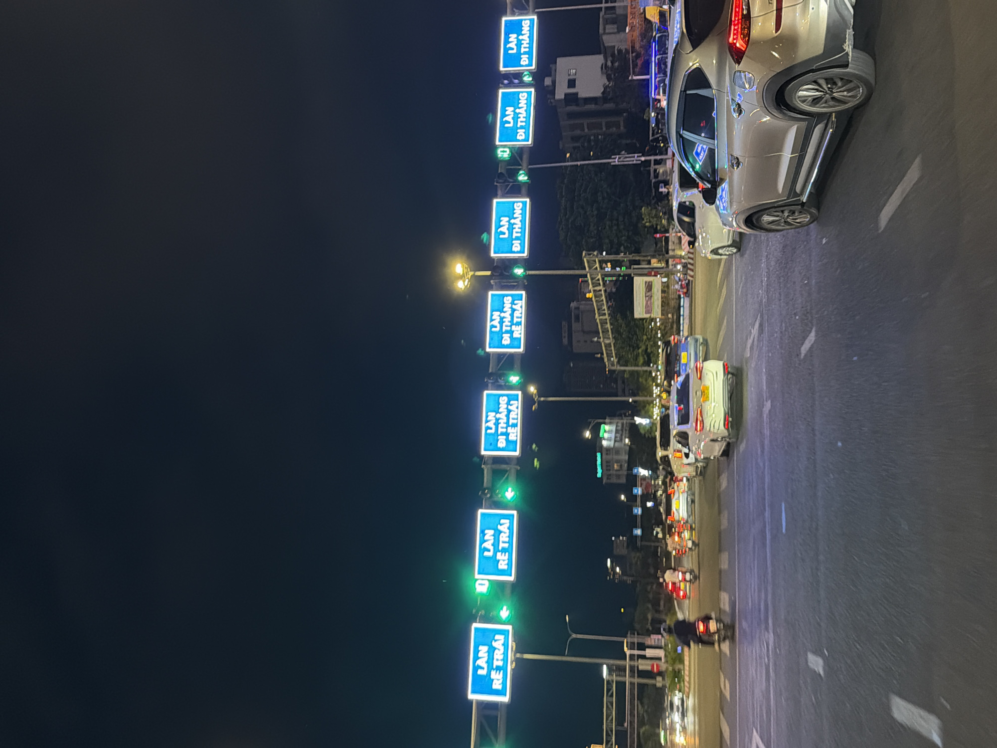 The image, taken at night, depicts a street in Đà Nẵng, Vietnam. The view is along a road with multiple lanes of traffic, where cars and motorcycles are visible. Overhead, there are numerous blue and white illuminated road signs with Vietnamese text indicating lane directions. The signs, stacked vertically, show arrows and traffic information for directions "straight" and "turn left." Streetlights and building lights provide illumination. The sky is dark, suggesting a late-night hour.