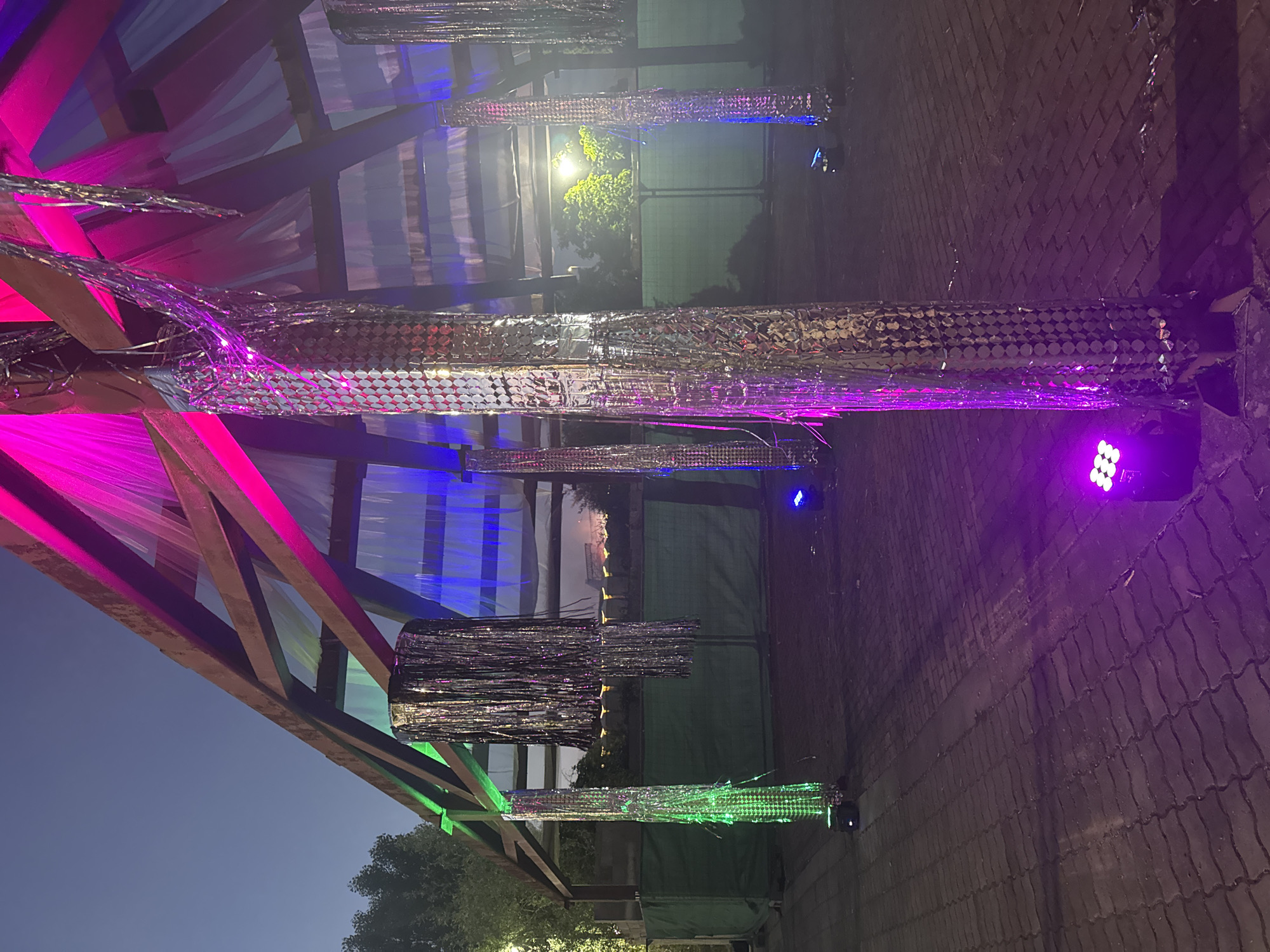 The image depicts an outdoor structure with a wooden frame, possibly a stage or canopy, decorated with silver tinsel streamers and fabric draping. The structure is illuminated by various colored LED lights, including vibrant pink, blue, and green, casting colorful reflections on the tinsel and surrounding pavement. Several tall, cylindrical objects covered in reflective material, resembling disco balls or sequined columns, are positioned within and around the structure. The ground is paved with interlocking bricks. In the background, there is a green fence and some trees, suggesting an urban or park-like setting. The sky is dark, indicating it is either dusk or nighttime. The overall scene conveys a festive or event-oriented atmosphere, likely a light installation or outdoor party setup. The location is specified as Sofia, Bulgaria.