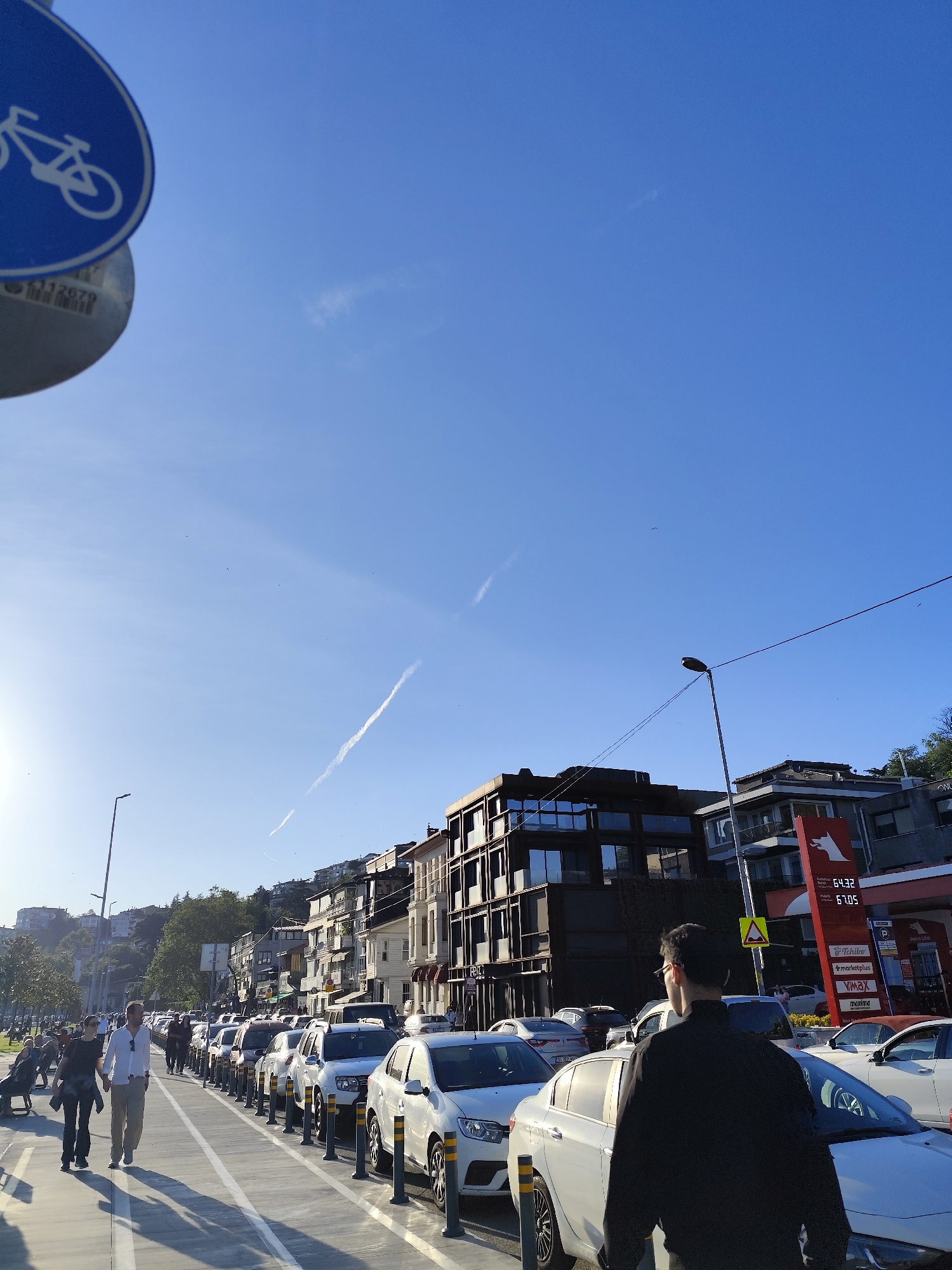The photograph captures a street scene in Sarıyer, Turkey, on a clear, sunny day. A blue bicycle path sign is visible in the upper left corner of the frame. The sky is a deep blue, with a prominent contrail streaking across it.
A row of parked white cars lines the right side of the street, separated from the pedestrian walkway by black bollards. Several pedestrians are walking along the walkway, including a man in a white shirt and khaki pants, and a person in black. A man in a black jacket and glasses stands near the foreground, partially obstructing the view.
Buildings of varying architectural styles line the street, including a modern dark-colored building with large windows and more traditional structures. A gas station with a red sign is visible further down the street. A light post is situated next to buildings.