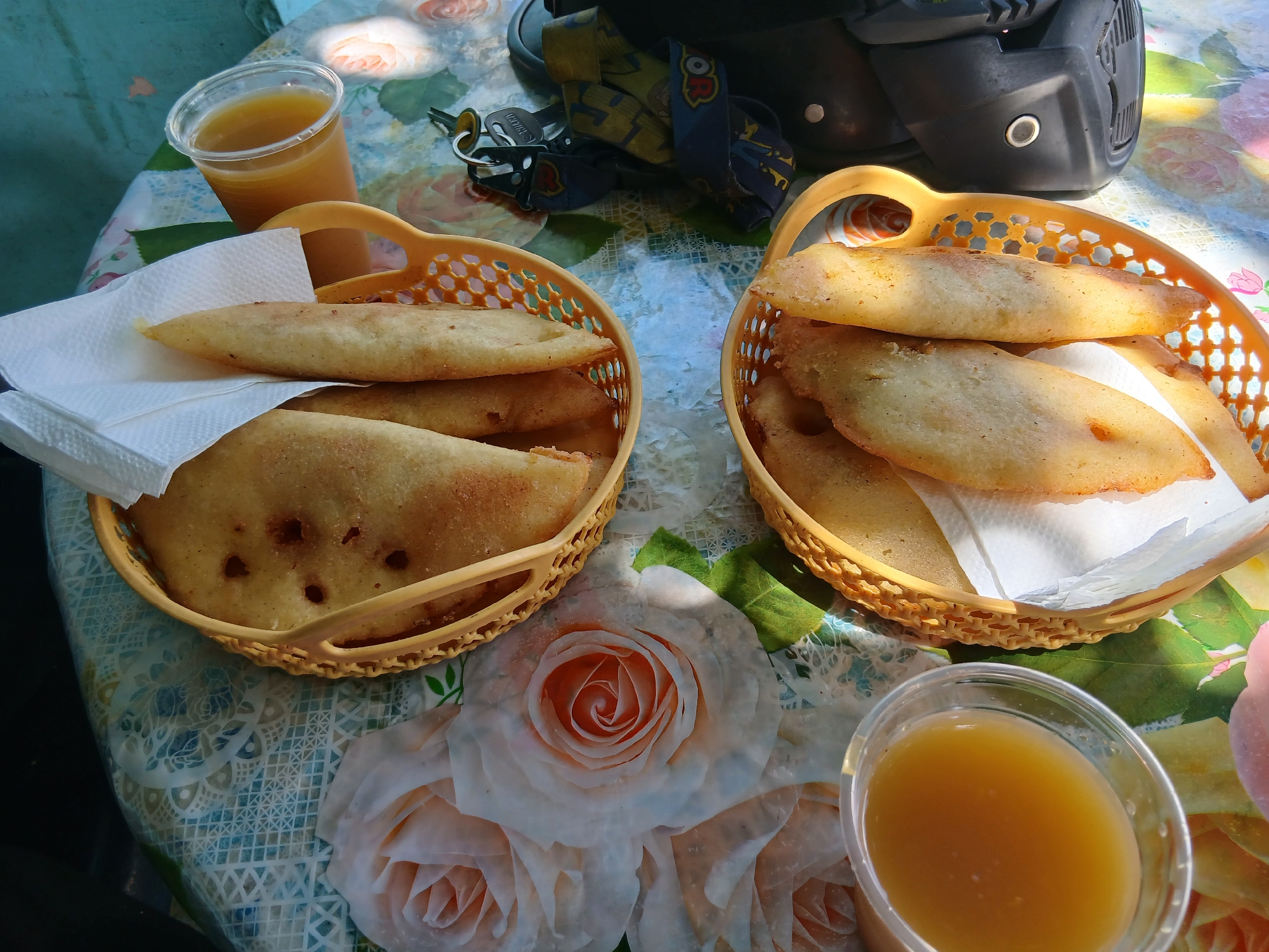 The image presents a table setting with Venezuelan cuisine, likely in or around Juan Griego, Venezuela, as implied by the prompt. Two woven baskets contain golden-brown empanadas, served on white paper napkins. Clear plastic cups of a yellow beverage, possibly juice, are situated near the baskets. A motorcycle helmet, keys, and lanyard lie on the table, suggesting the owner is nearby. The table is covered with a patterned tablecloth, primarily white, blue, and green, adorned with floral motifs of white and peach-colored roses. The scene appears to be outdoors in daylight.