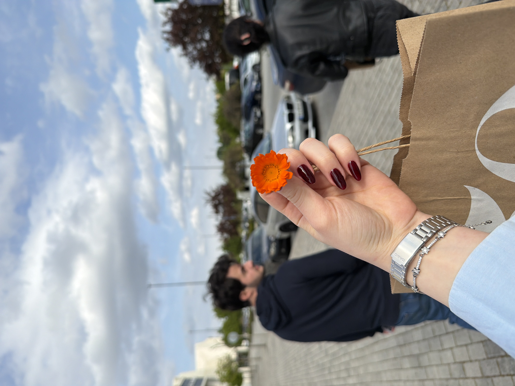 The image, rotated ninety degrees, captures a person's hand holding a vibrant orange flower (possibly calendula) against an outdoor backdrop. The hand features dark red, almond-shaped nail polish. On the wrist, a silver-toned watch with a metal link band is prominent, with "CASIO" visible on its clasp. Adjacent to the watch, a delicate silver bracelet adorned with star-shaped charms, each containing small white stones, is worn.
To the right, a brown paper shopping bag with rope handles is partially held, revealing a segment of a large, stylized white letter or logo. The background is out of focus but shows a paved parking area. Two other individuals are present: one closer, seen from behind, wearing a black leather jacket, and another further back, wearing a dark hoodie and seen from the side. Several cars, some with distinctive grilles suggesting BMWs, are parked along the area. Trees and distant buildings complete the scene under a cloudy, bright sky, indicating daytime. The setting appears to be an urban or suburban area, consistent with a shopping experience in Tbilisi, Georgia.