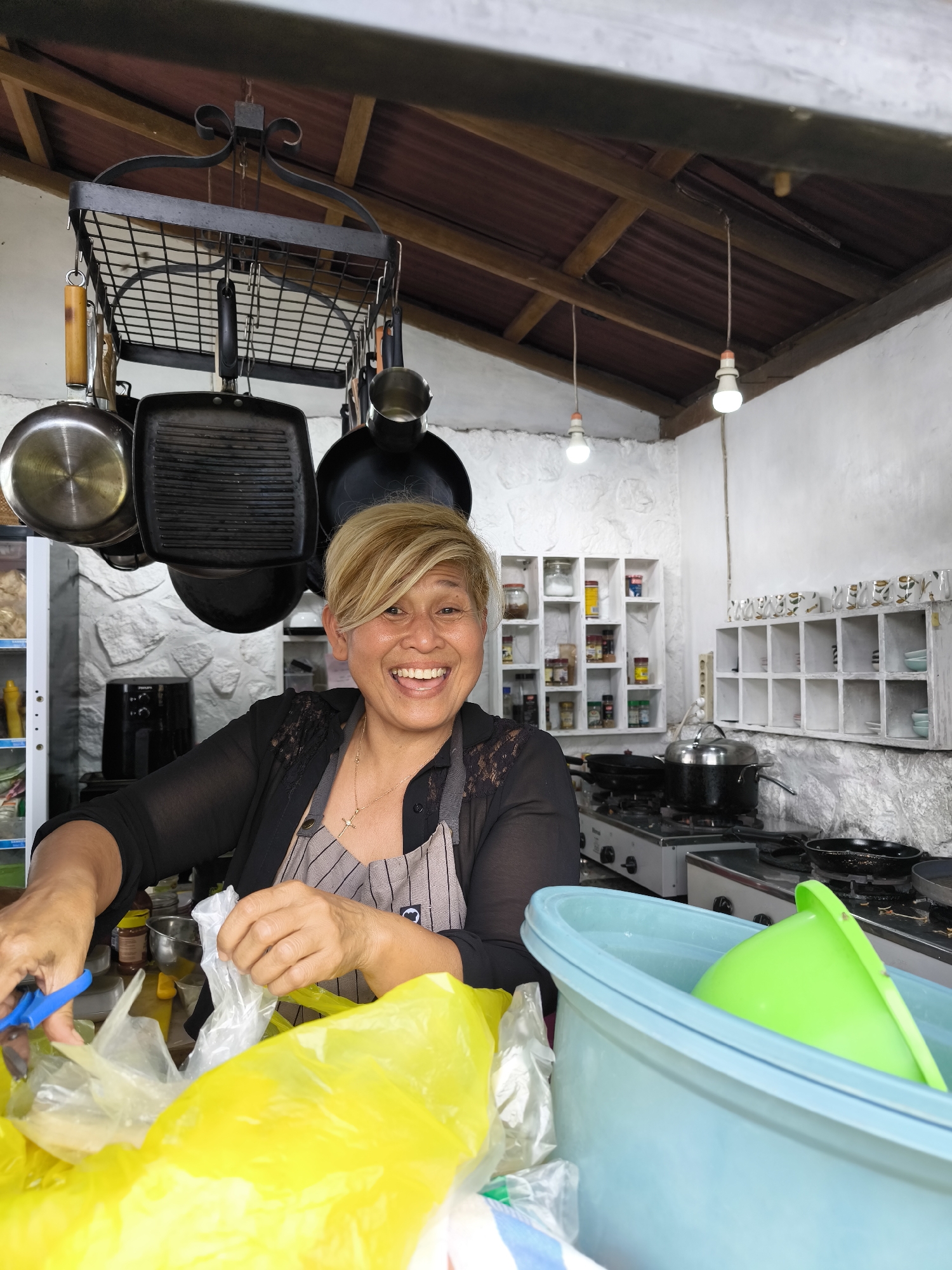 The image shows a woman with short blonde hair, smiling and cutting open a plastic bag with blue scissors in a kitchen setting. She is wearing a striped apron over a black top and a gold necklace with a cross pendant. Above her head, a rack holds various pots and pans. Behind her, a white-painted wall features shelves with containers and various items. A gas stove and a kettle are visible in the background. In the foreground, there is a blue tub filled with a green funnel. The location is unspecified, indicated by Unknown, Unknown.