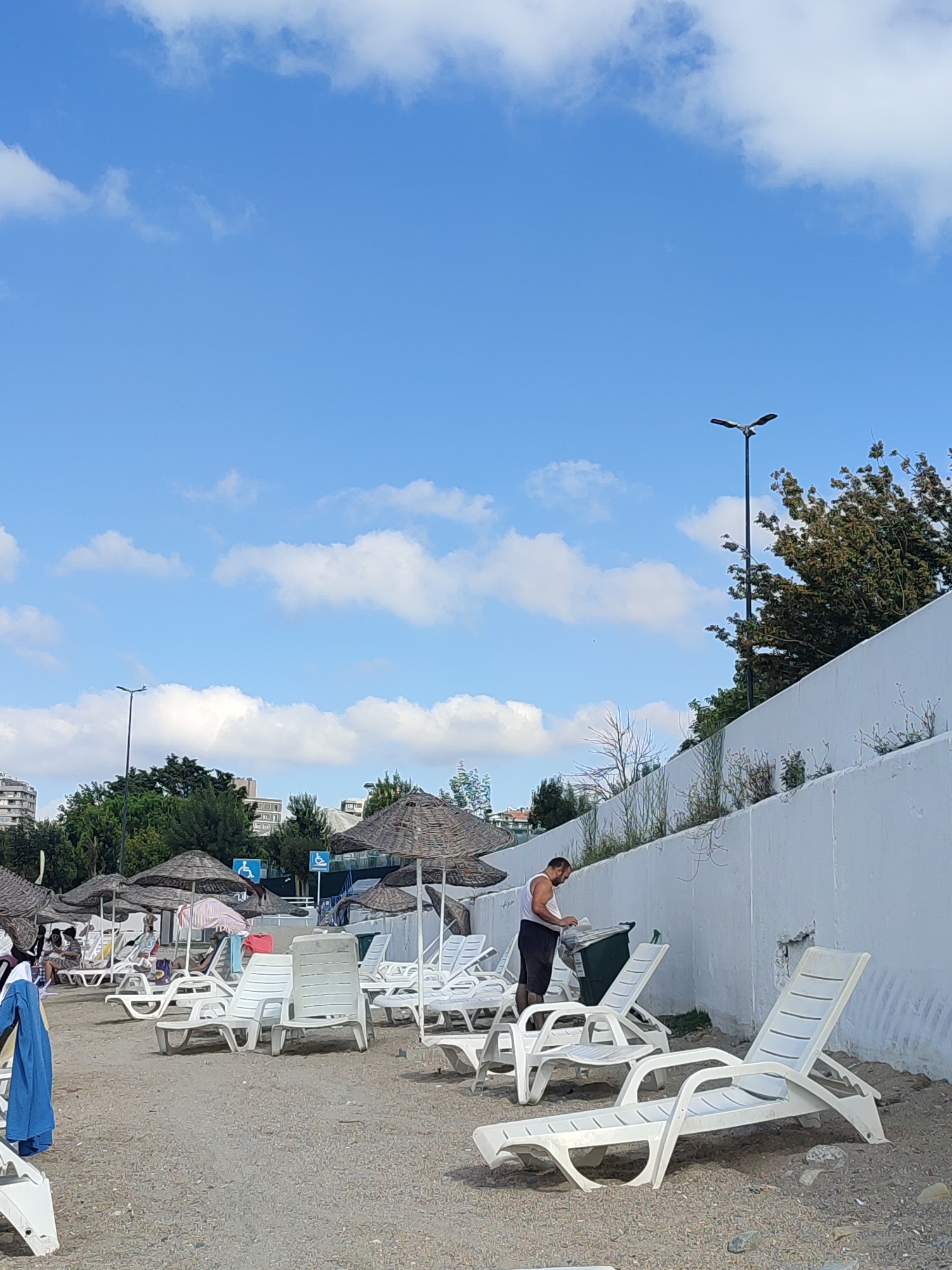 The image depicts a beach scene on a sunny day in Kadıköy, Turkey. In the foreground, white plastic lounge chairs are arranged on a sandy beach. Some are empty, while others have towels or bags on them, suggesting their use by beachgoers. Further back, more lounge chairs and thatched umbrellas are visible, with a few people seated under them. A man in a white tank top and dark shorts is seen standing near a green trash bin, appearing to be disposing of something. In the background, a white concrete wall runs along the edge of the beach, topped with greenery and trees. Buildings and a street light are visible beyond the wall, indicating an urban setting. The sky is blue with scattered white clouds.