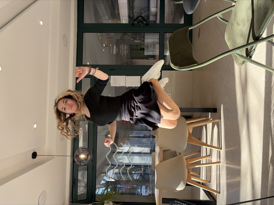 An adult female is captured mid-jump within a café interior, identified as 'More' by reversed signage visible on its glass facade in Budva, Montenegro. The woman, with long, wavy blonde hair appearing windblown, wears a black long-sleeved top, a black flared mini-skirt, and white sneakers. Her knees are bent, and arms are slightly raised, one with an open palm and the other a loose fist.
The café features a light-colored tiled floor with prominent shadows cast by direct sunlight entering through a large glass facade with dark green frames. The white ceiling contains multiple recessed spotlights and a single spherical grey pendant lamp. To the left, a light wood table with black metal legs is surrounded by beige chairs with wooden bases. A dark olive green upholstered folding chair occupies the right foreground. A small hanging plant is visible near the left side of the glass facade.
Through the glass facade, an urban street scene is visible, including parked vehicles, trees, and distant buildings. Reflected text on the glass includes words like 'LOVE', 'COFFEE', and 'DAY' along with the mirrored 'MORE' café name.