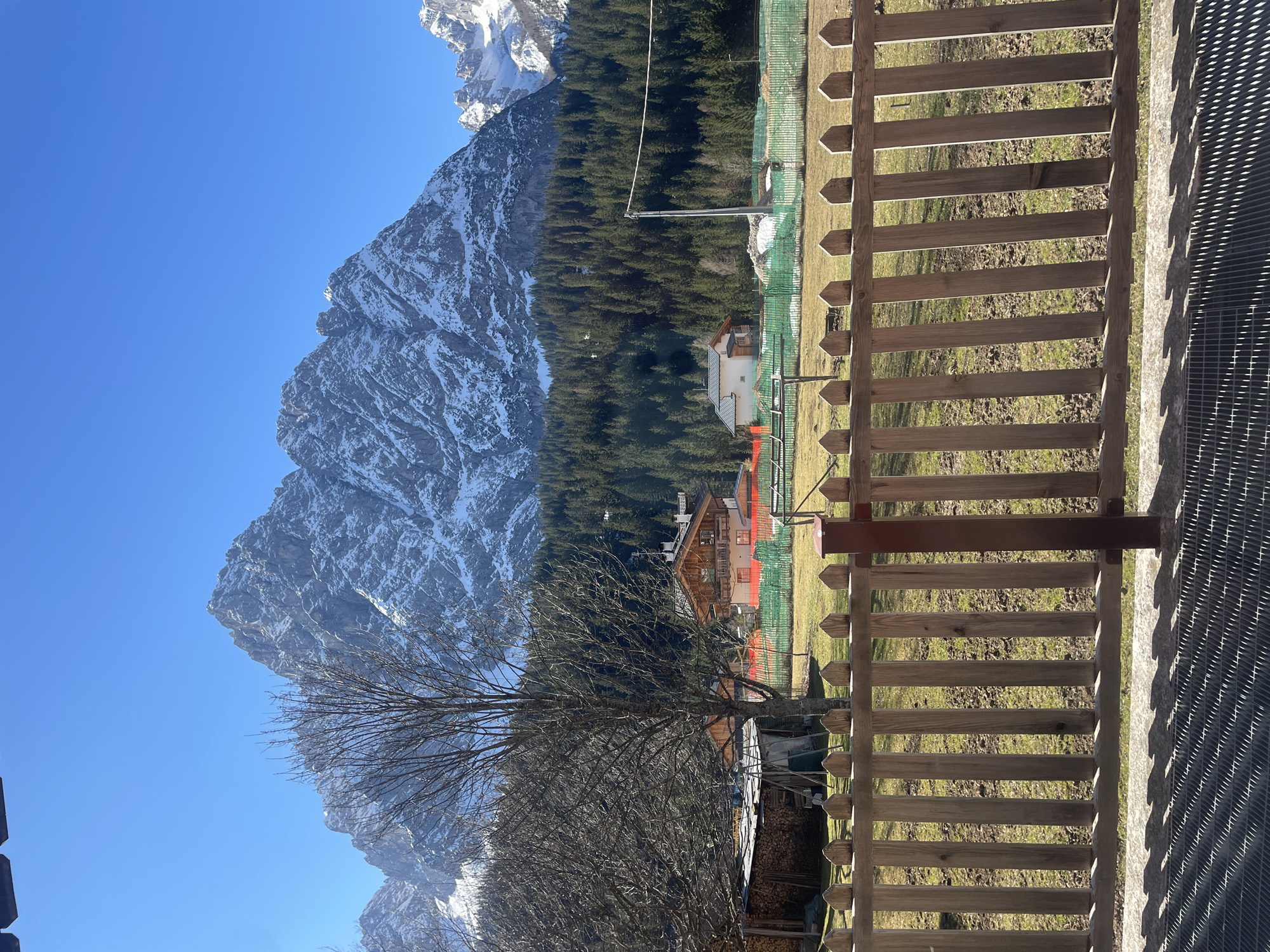 The image shows a landscape in Sappada, Italy, featuring a prominent mountain partially covered in snow under a clear blue sky. A dense evergreen forest extends from the base of the mountain towards the foreground. A wooden fence runs vertically across the right side of the frame, partially obscuring a grassy field. In the middle ground, a cluster of buildings can be seen amongst the trees, including structures with wooden architecture and orange safety netting around them. A bare tree is visible in front of the buildings. In the lower right corner of the frame, a metal grate surface is visible. The photo has been taken with a slight tilt.