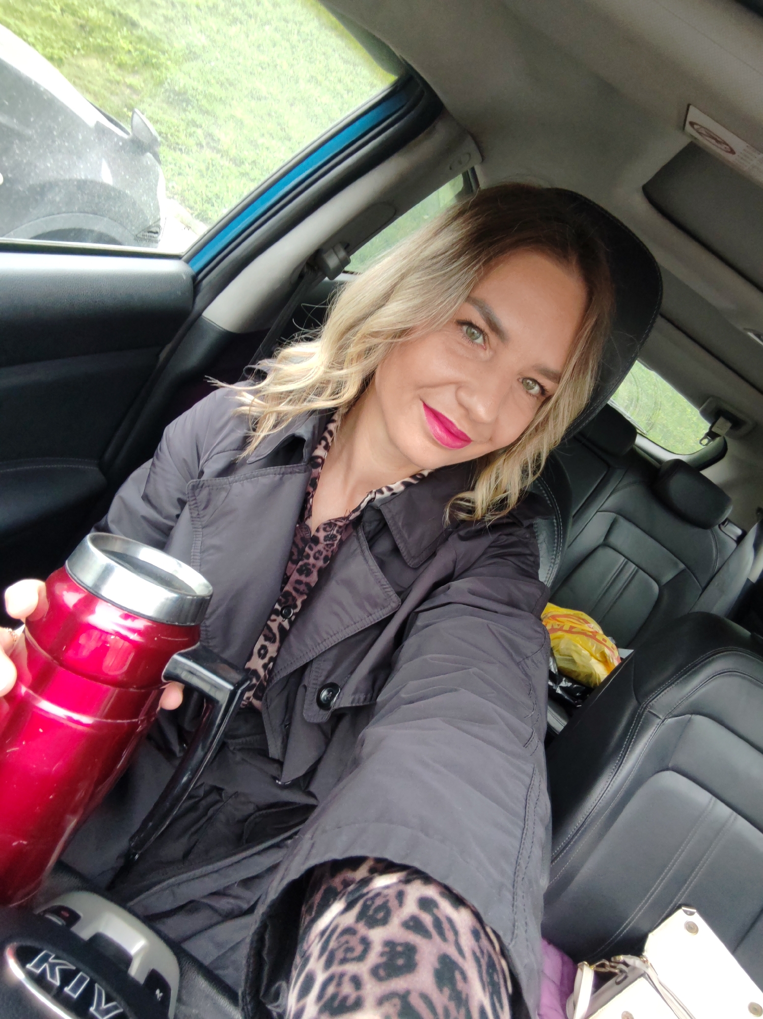 A woman with blonde, wavy hair and green eyes is taking a selfie inside a car. She has bright pink lipstick and a slight smile. She is wearing a dark grey or black trench coat-style jacket over a leopard print top or dress, parts of which are visible on her chest and leg. In her left hand, she holds a shiny red thermos or travel mug with a silver rim and a black handle.
The car's interior features dark leather seats. A yellow plastic bag is visible on the front passenger seat, and a white object, possibly a purse, is on the floor or seat next to it. Through the windows, green foliage and what appears to be another dark-colored car are visible, suggesting an outdoor, possibly natural, setting. The lighting seems to be natural daylight, possibly overcast. A blue trim is visible along the car's interior frame near the window. Part of the car's gear selector or console is visible in the foreground, with the letters "KIV" or "KIA" partially inscribed on it, likely indicating the car's brand. The overall scene suggests a casual moment during transit.