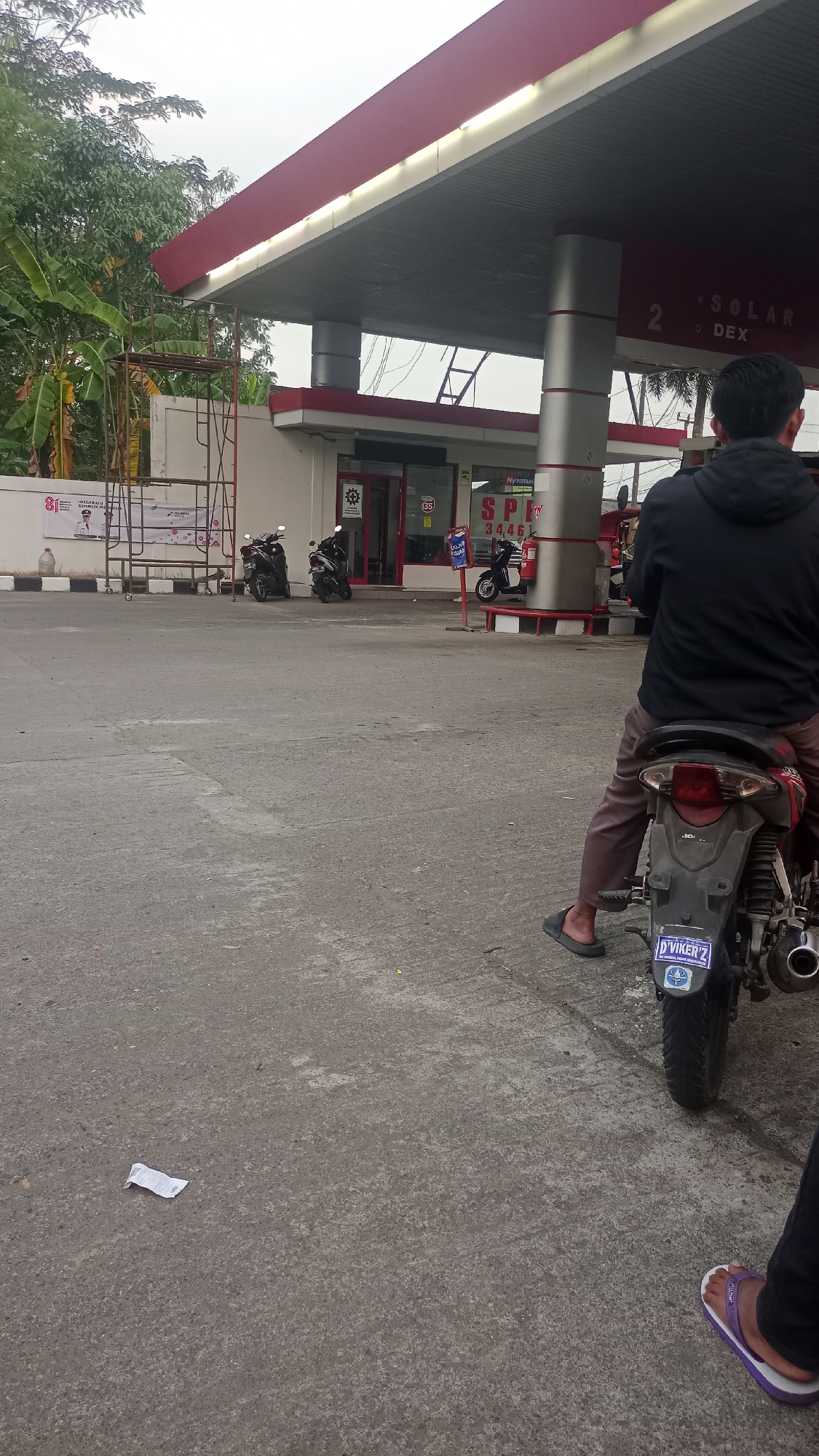 The image captures an outdoor scene at a gas station in Cogreg, Indonesia. In the foreground, a person is seated on a motorcycle, facing away from the camera. The motorcycle is black and red, with a license plate that reads "D'VIKER'Z". The person is wearing brown pants and black flip-flops. To the right, another person's legs and feet are visible, wearing purple flip-flops.
The gas station features a red and white awning supported by gray pillars. Signs on the pillars indicate "2 SOLAR DEX". The station building has an entrance with a red door and windows. Several motorcycles are parked in front of the building. To the left, a banner with Indonesian text and imagery is displayed on a white wall.
The ground is paved with concrete, and a crumpled piece of paper is visible on the ground in the lower left portion of the image. Trees and foliage are present in the background, suggesting an outdoor environment. The sky is overcast. The scene appears to be a typical day at a local gas station.