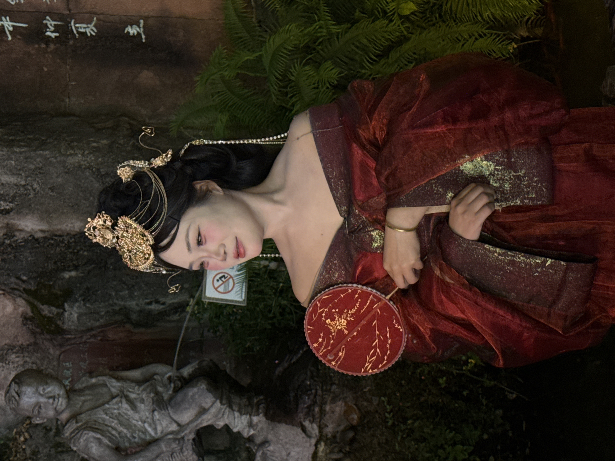 A woman in traditional Chinese attire stands outdoors in what appears to be a park or garden setting, possibly in the Yuzhong District of China, given the presence of a stone statue resembling a child and Chinese calligraphy on a wall. The woman is adorned with an elaborate golden headdress and wears a flowing, deep red gown with gold embellishments. She holds a circular red fan with a gold design in her left hand, and her right hand rests on her torso. Her makeup features prominent pink blush. In the background, a stone statue of a boy is visible, and to the right, lush green ferns and a wall with Chinese characters contribute to the environment. A small sign with a prohibition symbol is also present.