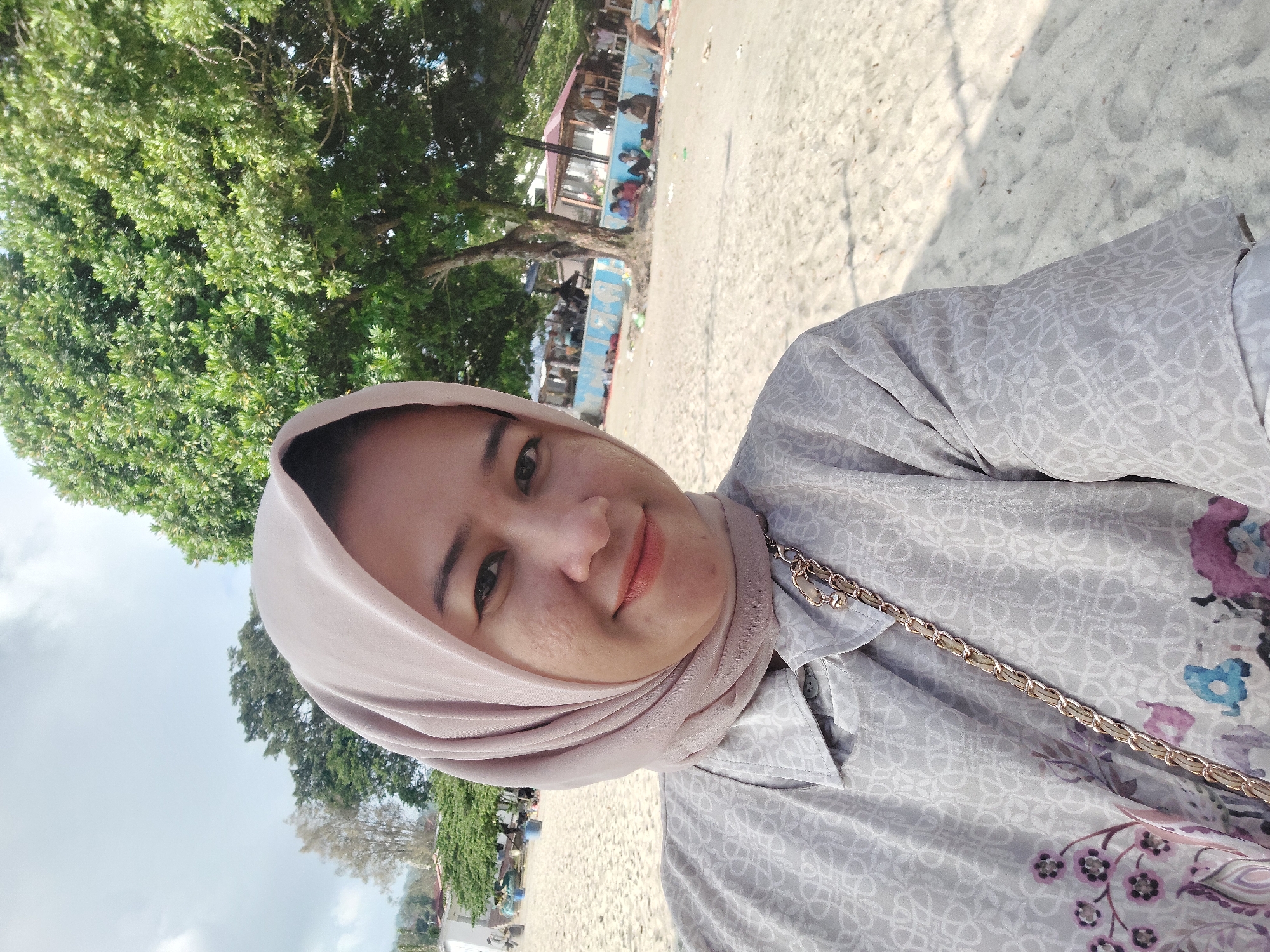 A medium shot captures a woman wearing a light pink hijab and a grey patterned shirt standing on a sandy beach. She is looking at the camera and smiling. Behind her, there is a large green tree, and further in the background, there are other trees, buildings, and people gathered. The sky is partly cloudy. The setting appears to be a beach in Parapat, Indonesia, with the distinct environment of a tropical location.